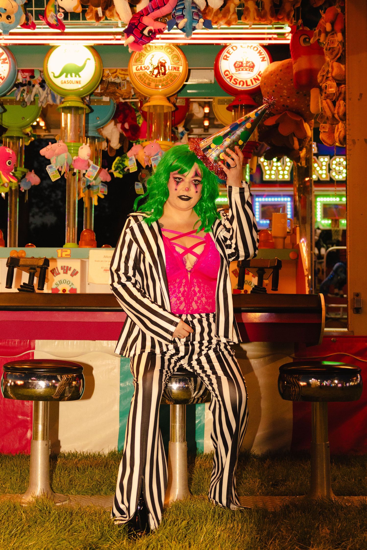 Costumed performer in striped suit and green wig posing at colorful carnival game booth.
