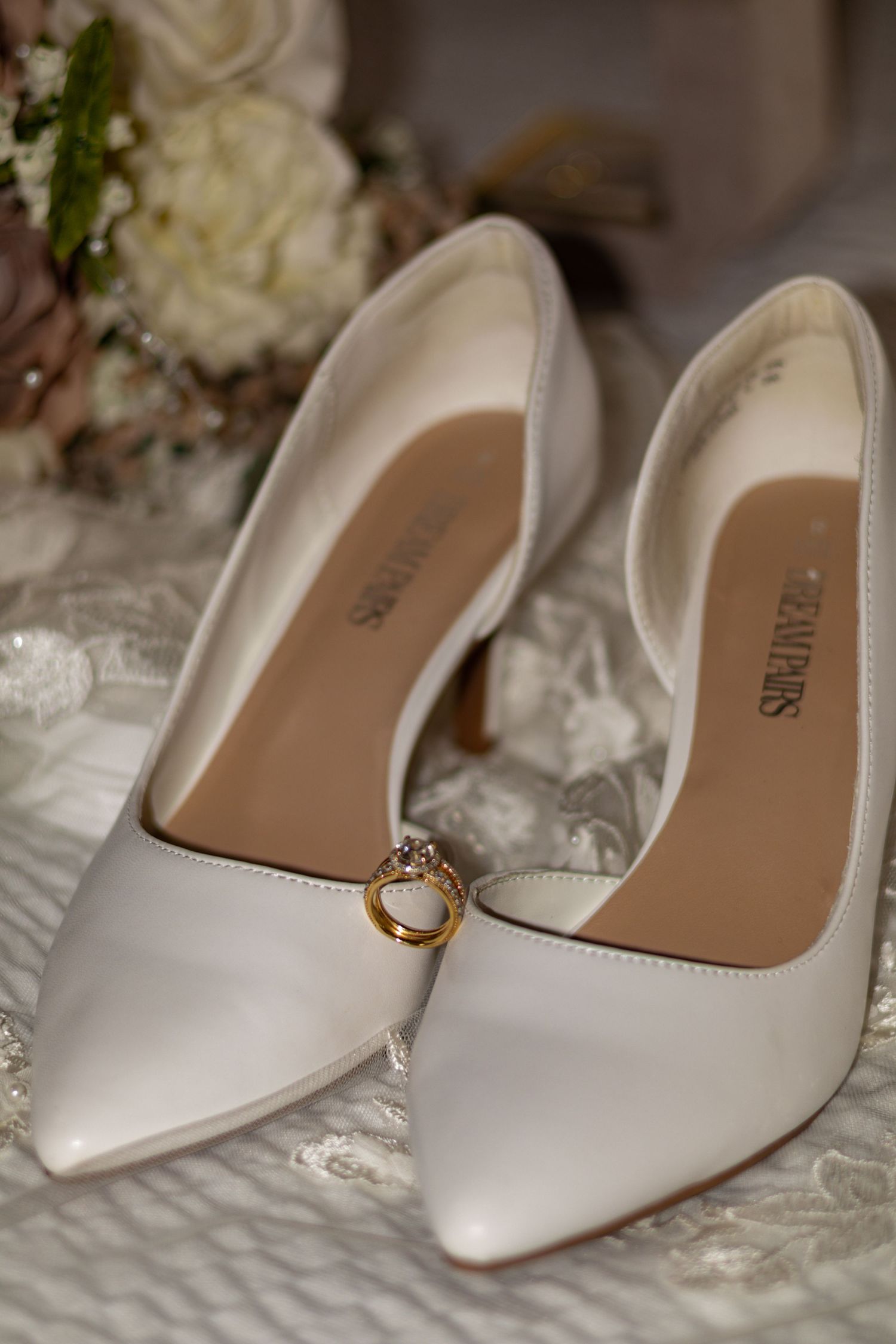 Elegant white bridal shoes with a delicate ring placed on lace fabric.