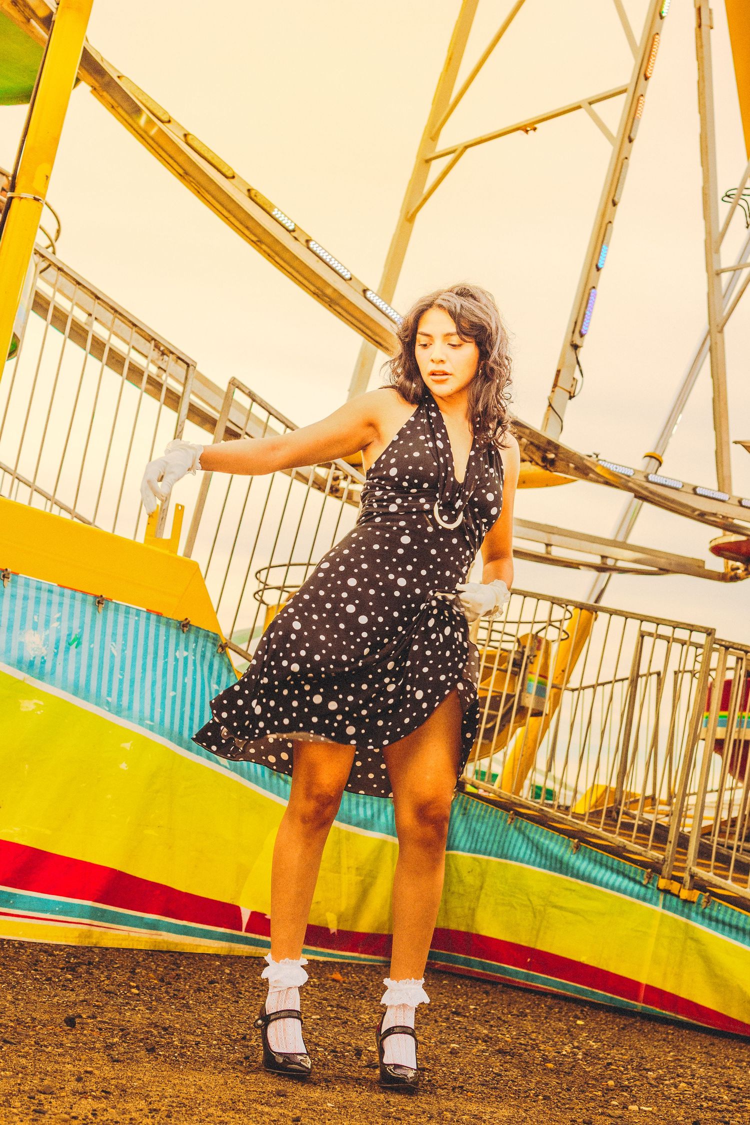 Young woman in black polka dot dress posing gracefully at a colorful fairground with a Ferris wheel backdrop.