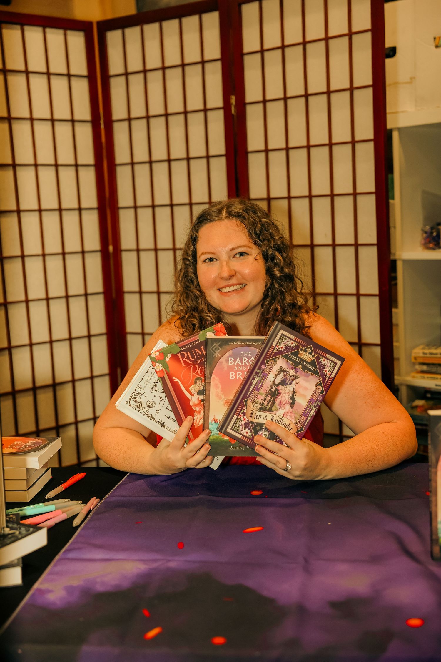 Smiling woman holding a fan of colorful books at a vendor table with purple cloth at an indoor book fair.