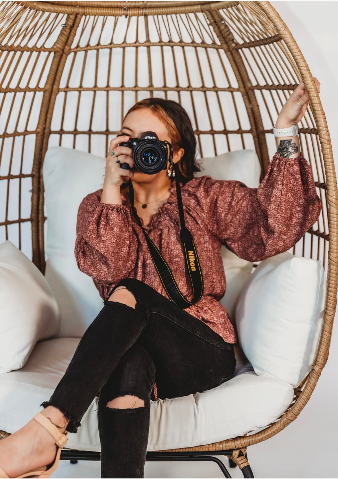 A photographer lounges in a rattan hanging chair wearing a pink patterned top and black pants while holding a camera.