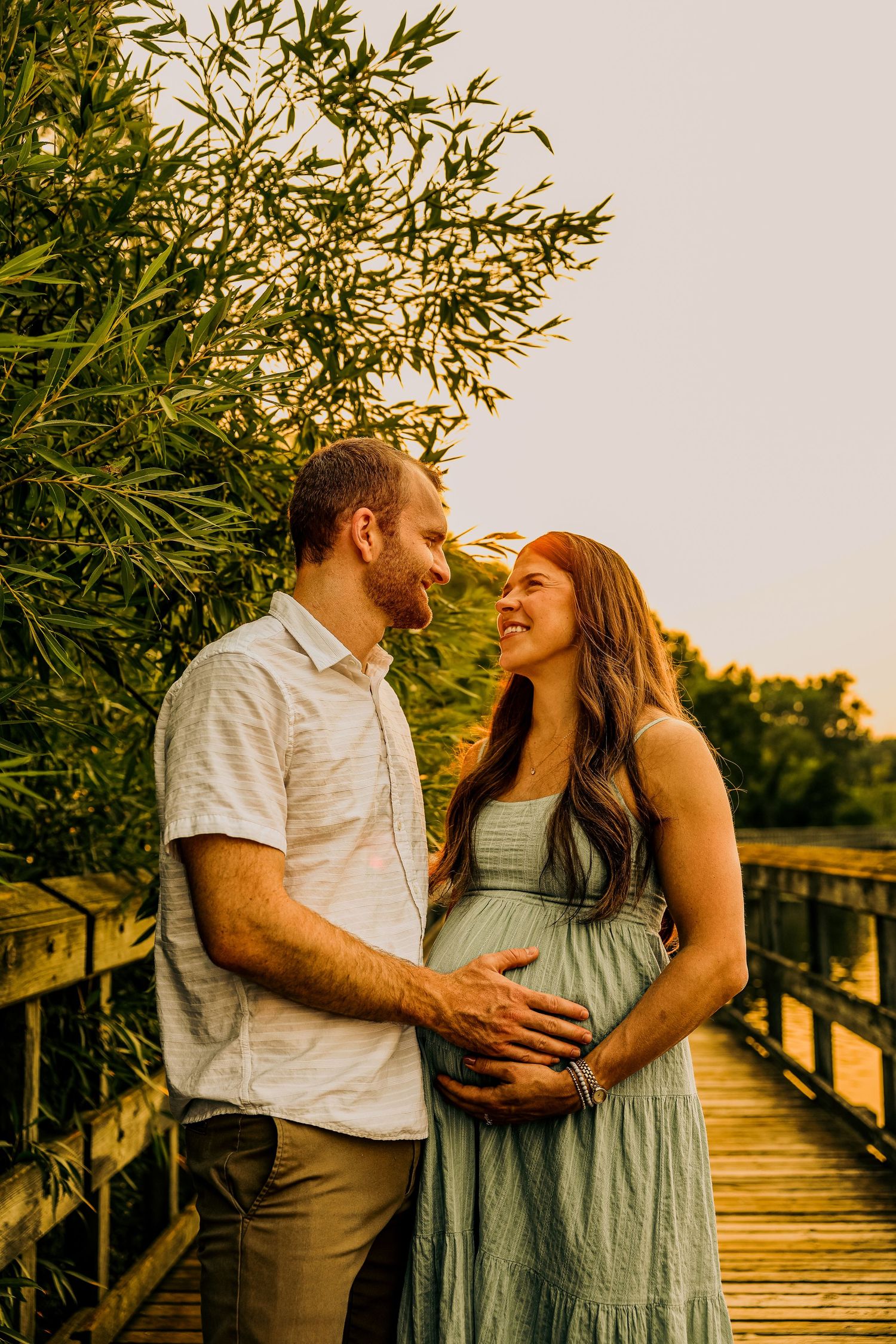 Couple sharing a tender moment on a wooden bridge at golden hour, with the woman in a sage green maternity dress.