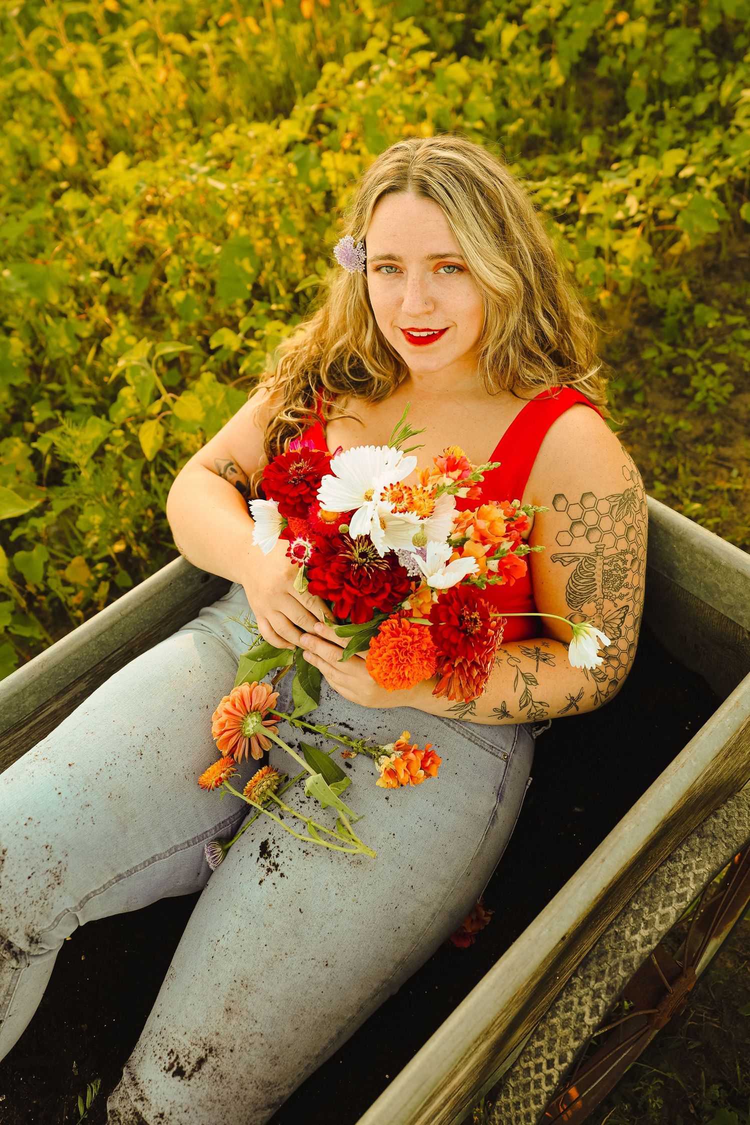 Woman with tattoos sitting in a wheelbarrow holding vibrant red and white flowers, surrounded by lush green foliage.
