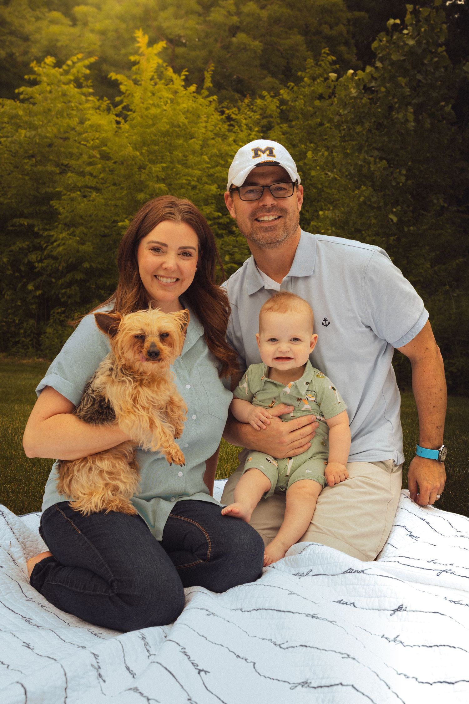 A smiling couple sits on a blanket outdoors, holding a baby and a small Yorkshire Terrier dog, with lush green trees behind them.