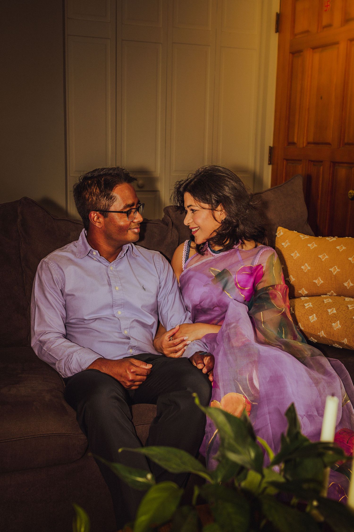 A couple sitting together on a couch, smiling at each other, woman wearing a purple saree in warm indoor lighting.