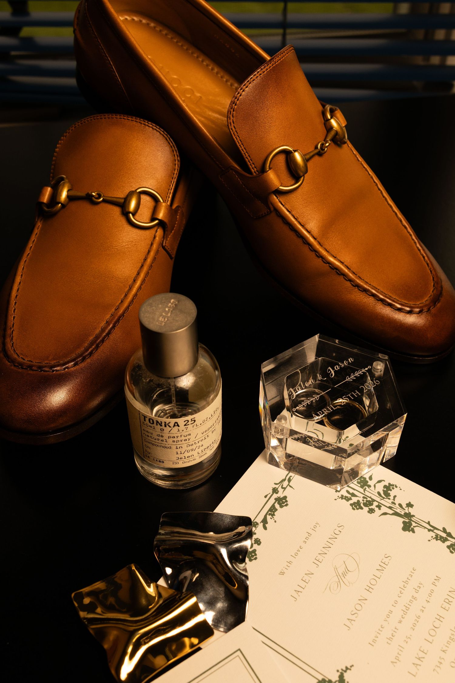 Brown leather Gucci horsebit loafers displayed with Le Labo fragrance, crystal perfume bottle, and wedding invitation.