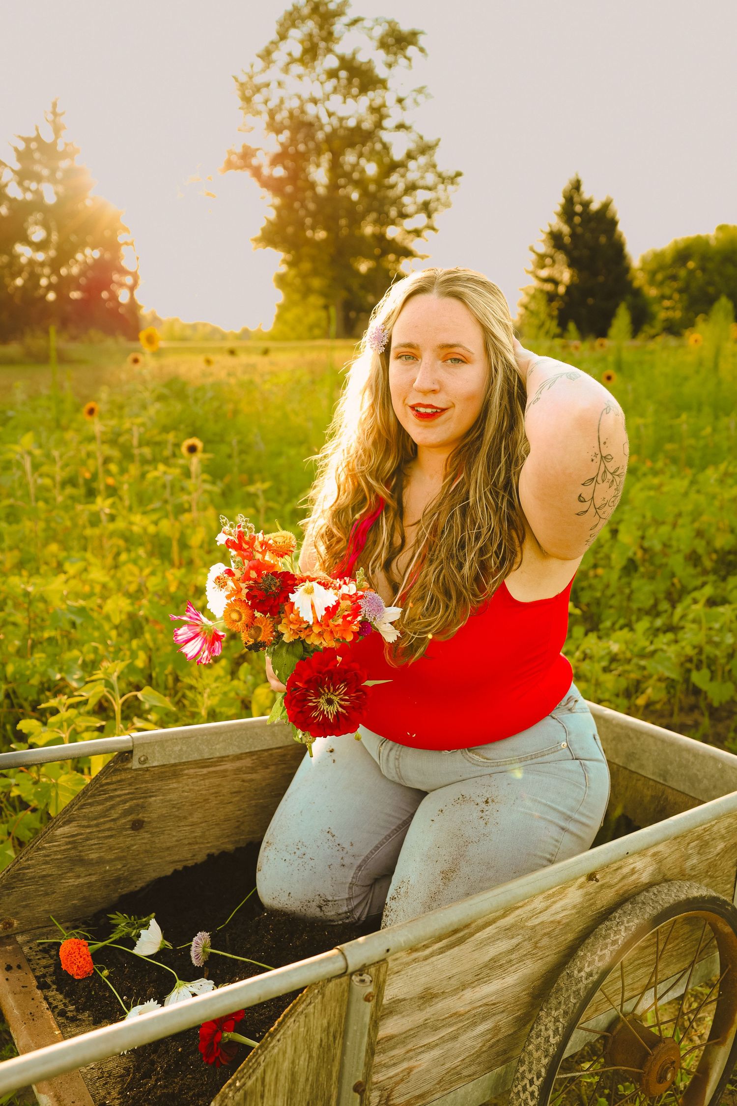 Woman in red top holding colorful flowers in a wooden wheelbarrow in a sunlit garden field.