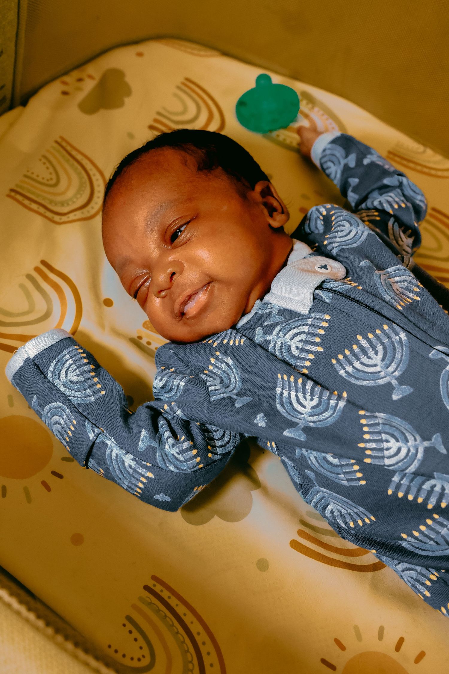 Smiling newborn baby in a blue Hanukkah menorah print onesie lying on a yellow patterned cushion.