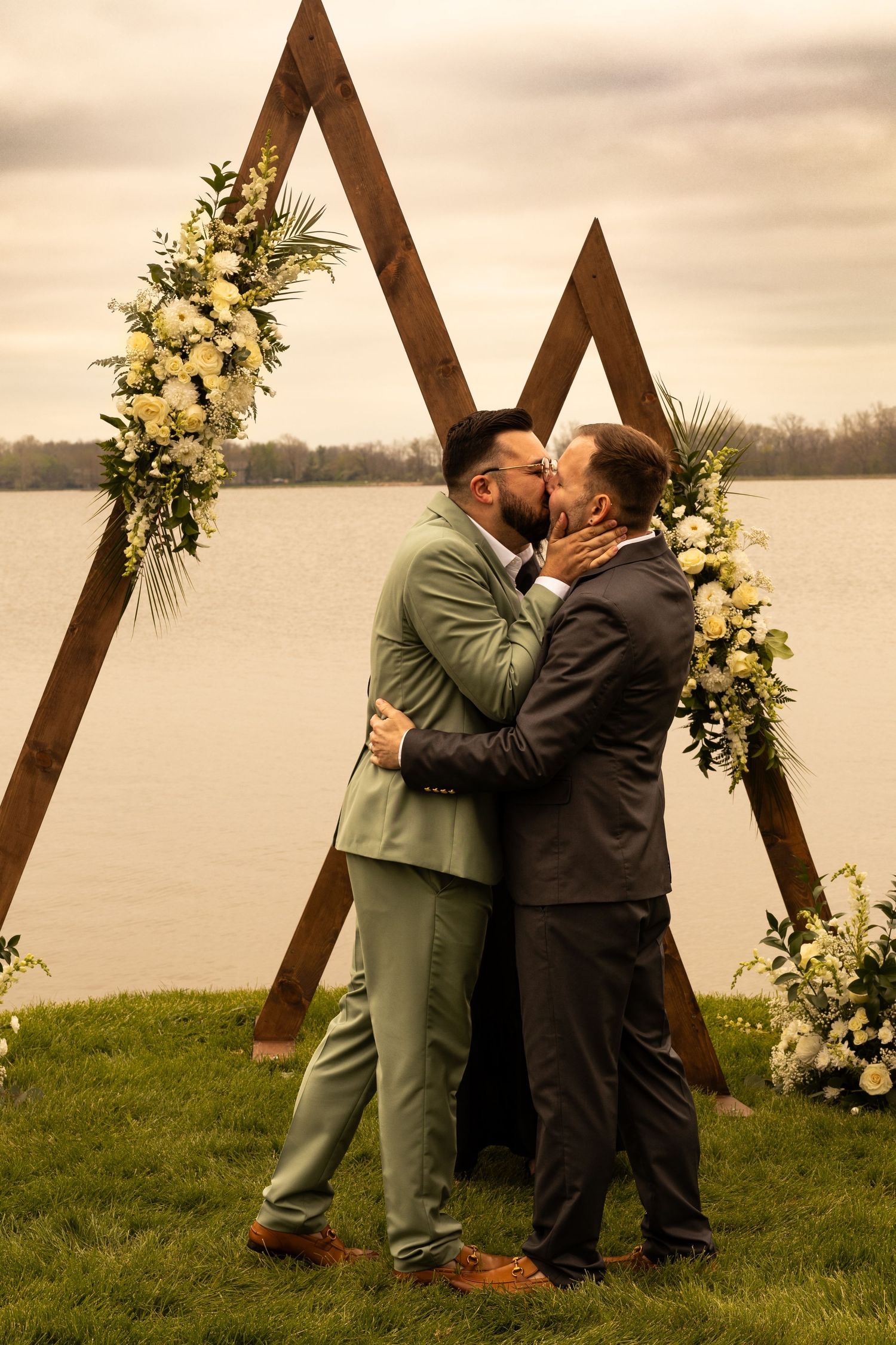Two grooms share a first kiss at outdoor wedding ceremony by a lake with a wooden triangular arch adorned with white flowers.