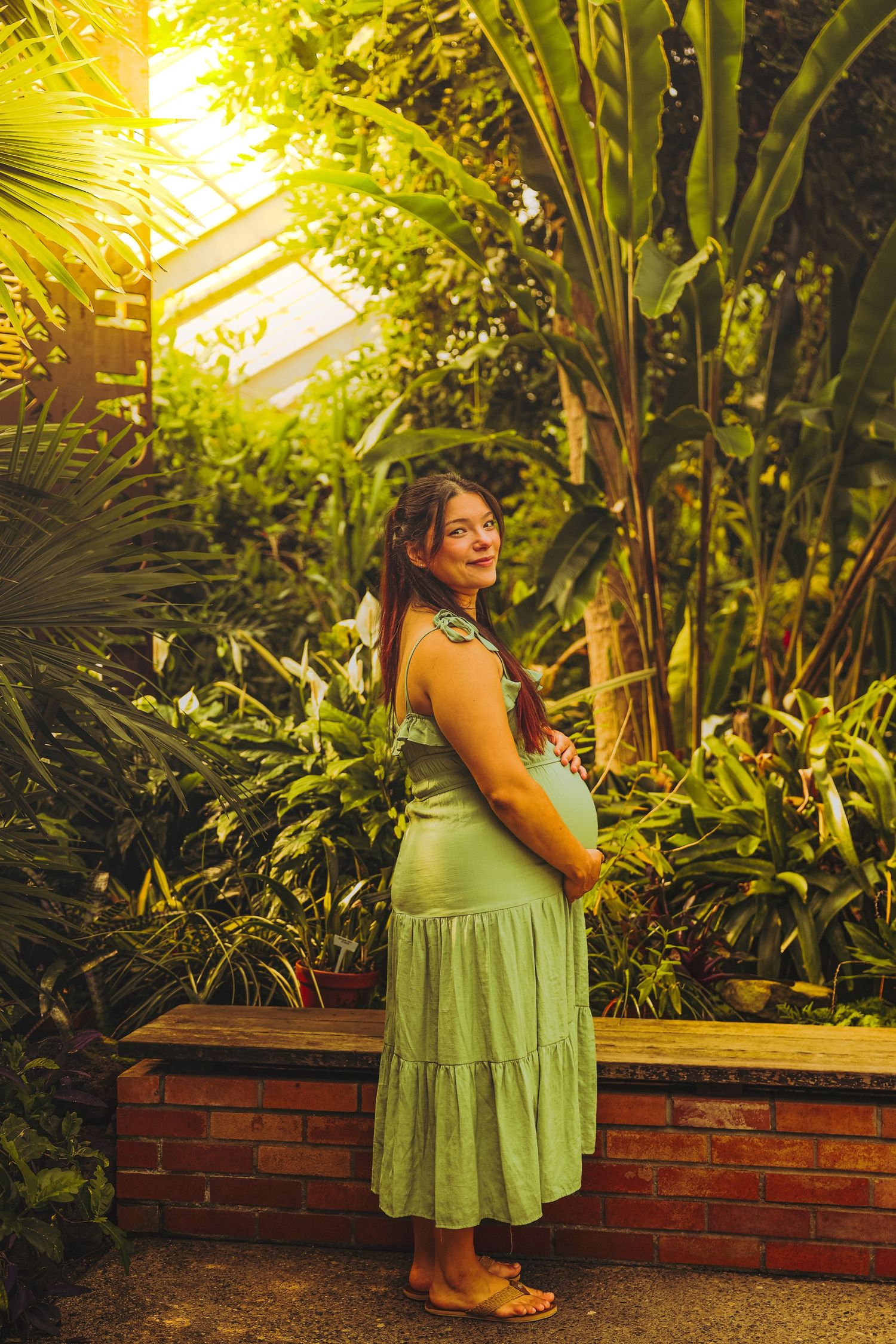 Pregnant woman in green dress posing in lush tropical greenhouse, cradling baby bump, surrounded by vibrant green plants.