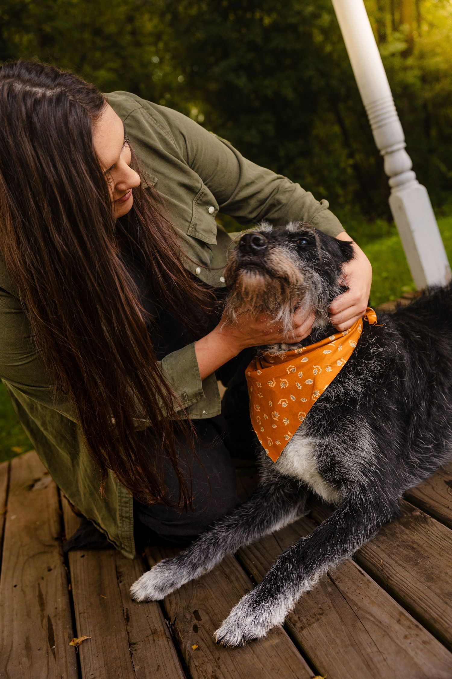 Woman in green jacket cuddling her schnauzer dog wearing an orange bandana on a wooden deck.