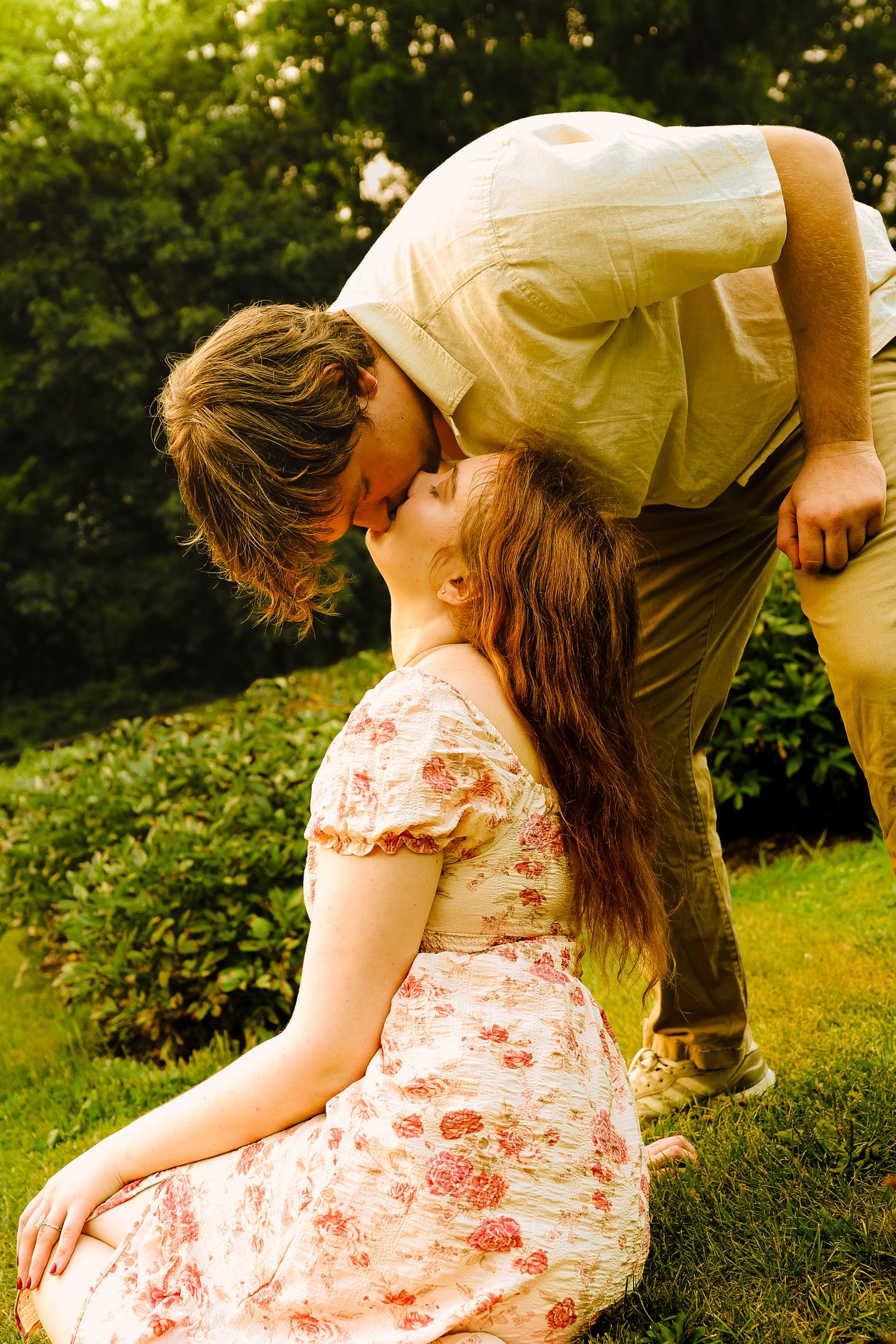A young couple shares a romantic kiss outdoors, with the man leaning down to kiss a woman in a floral dress.