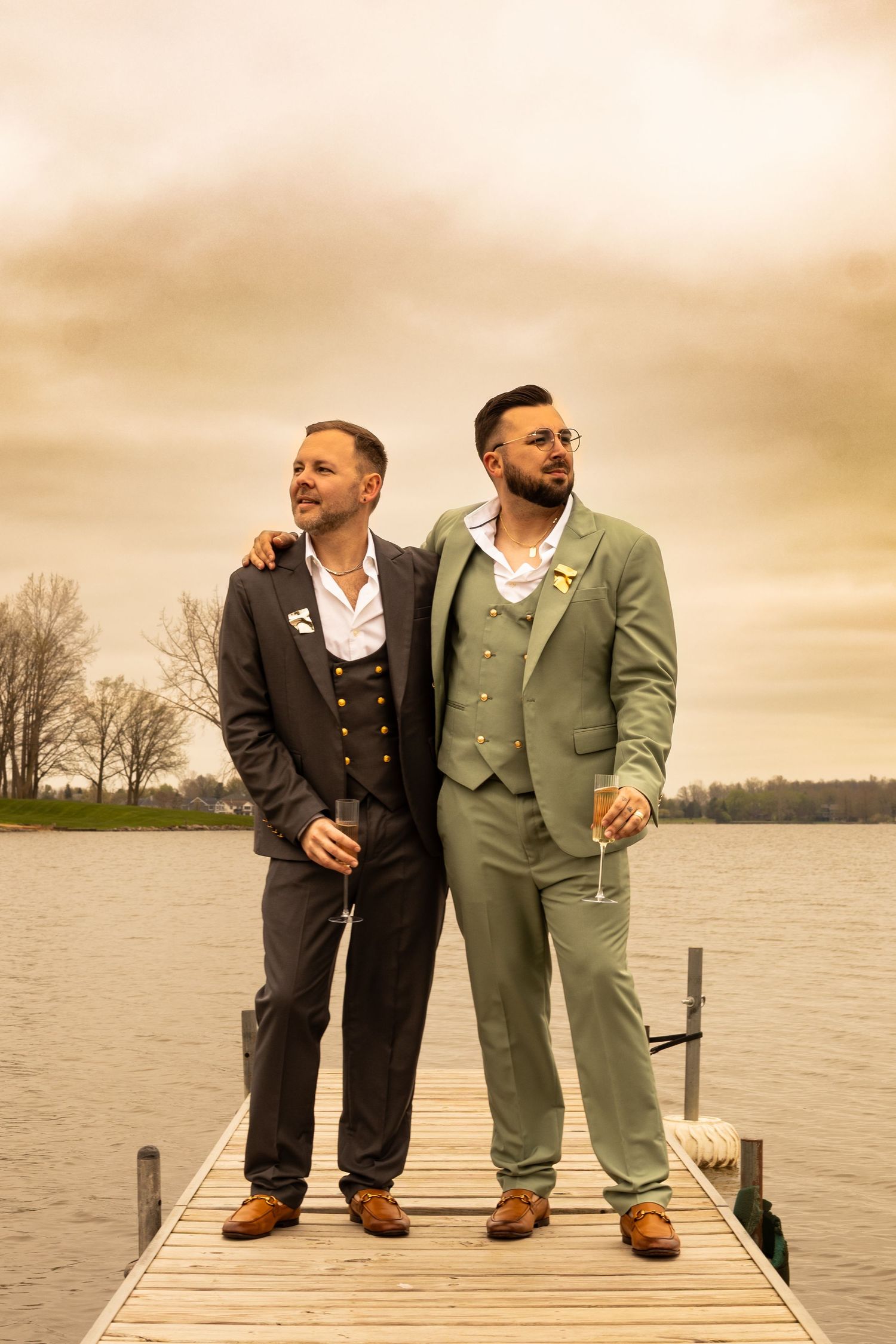 Two grooms in suits holding champagne glasses on a dock by a lake on their wedding day.