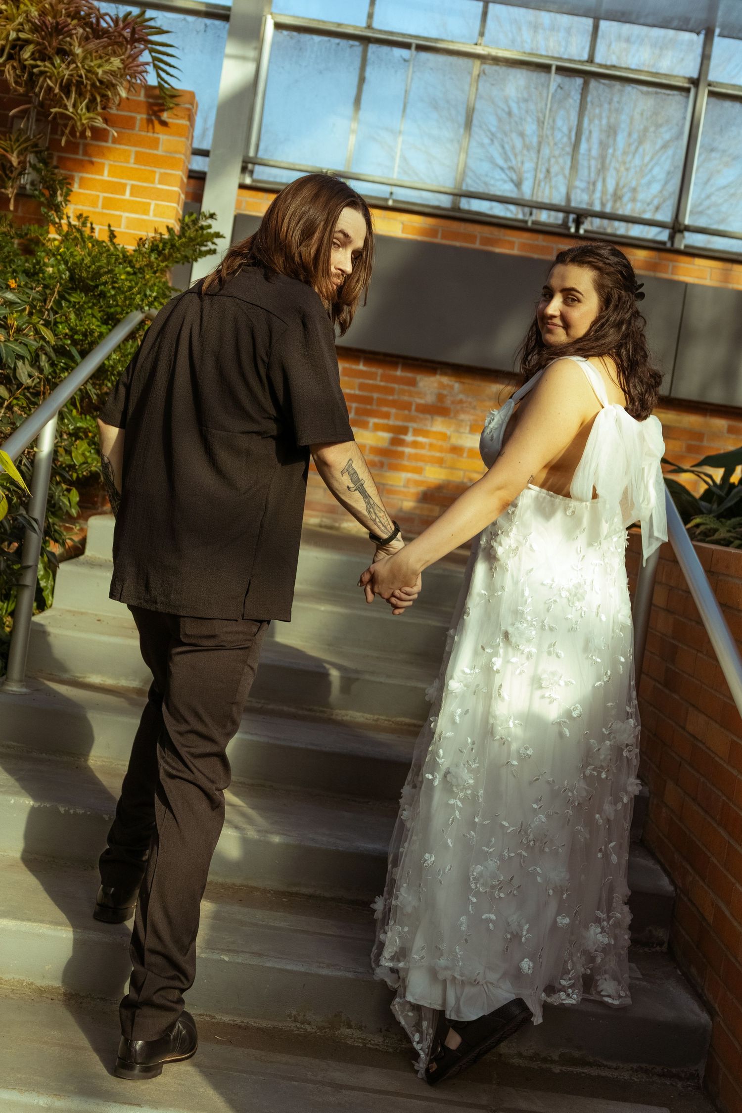Bride in white floral gown and groom in black suit hold hands walking up outdoor stairs on their wedding day.