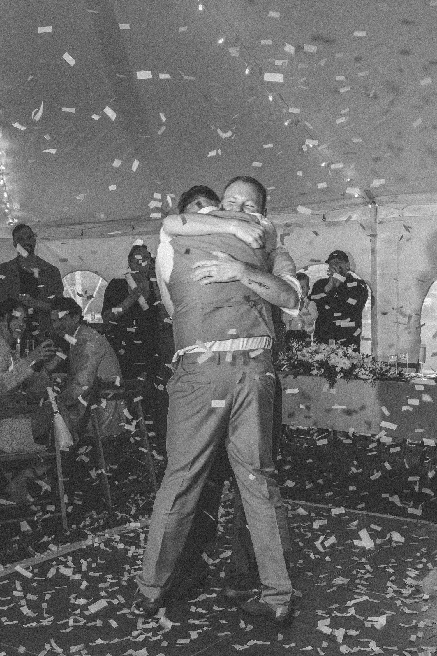 Two grooms share a joyful embrace on the dance floor surrounded by falling confetti at their wedding reception.