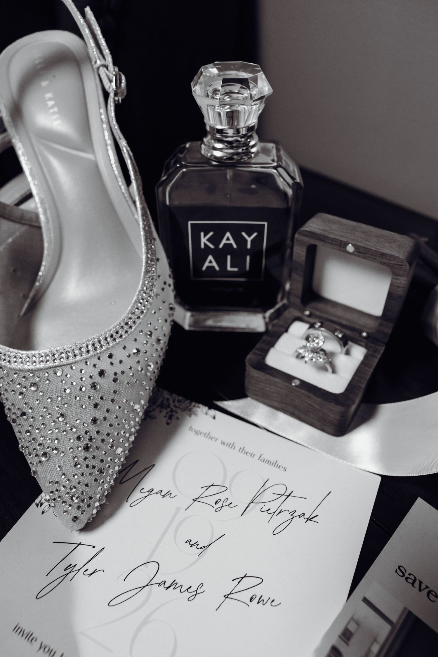 Black and white wedding flat lay with a sparkly slingback heel, Kayali perfume, diamond rings in a wooden box, and a printed invitation.