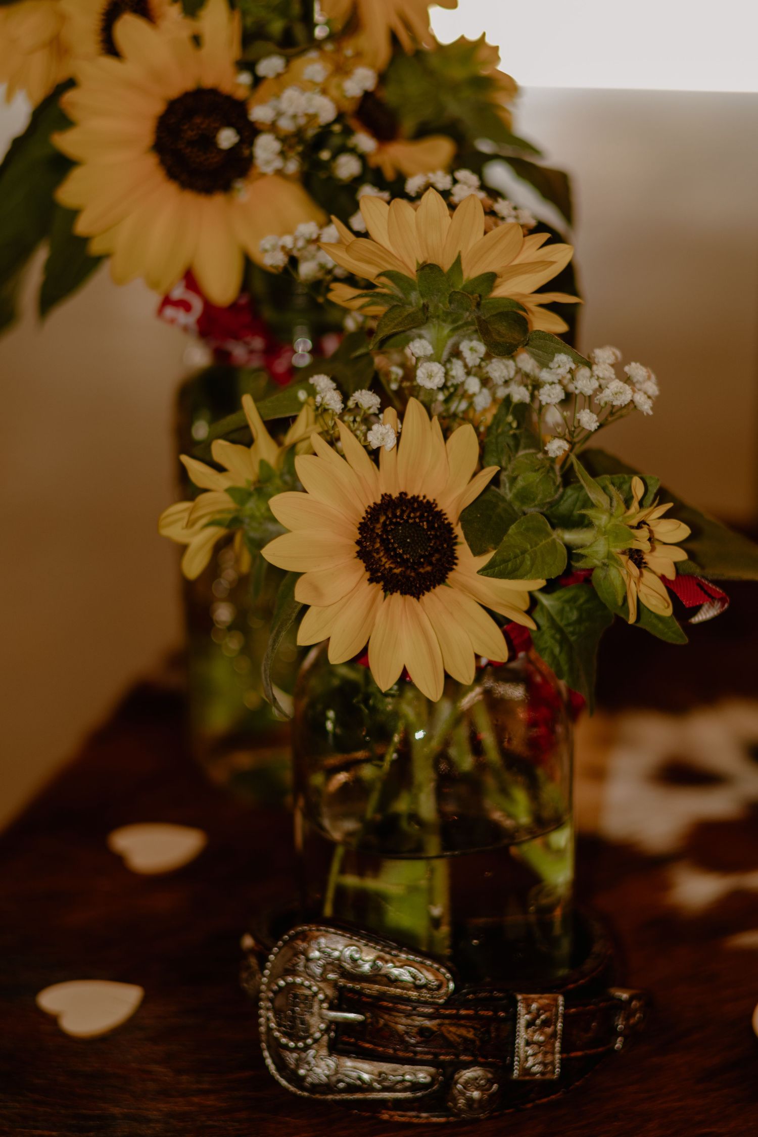 A rustic arrangement of yellow sunflowers and baby's breath in a glass jar sits on a dark wooden table.