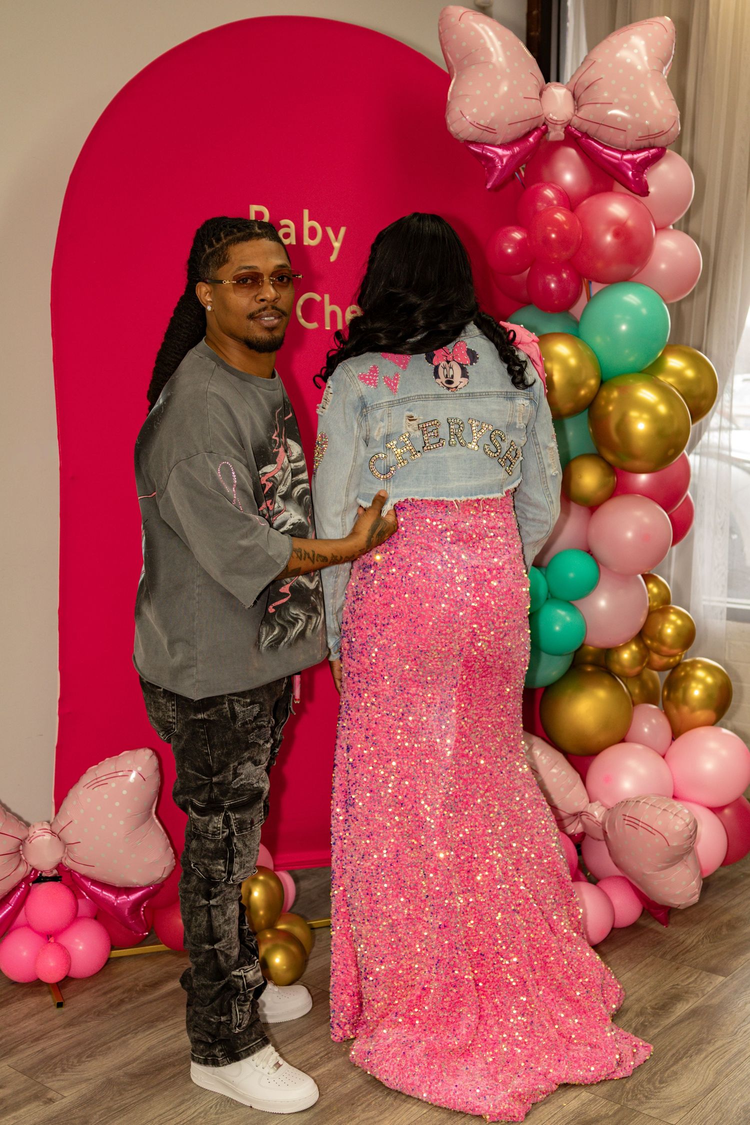 Couple posing at a pink Minnie Mouse baby shower with balloon arch, woman in sparkly pink maternity gown and denim jacket.
