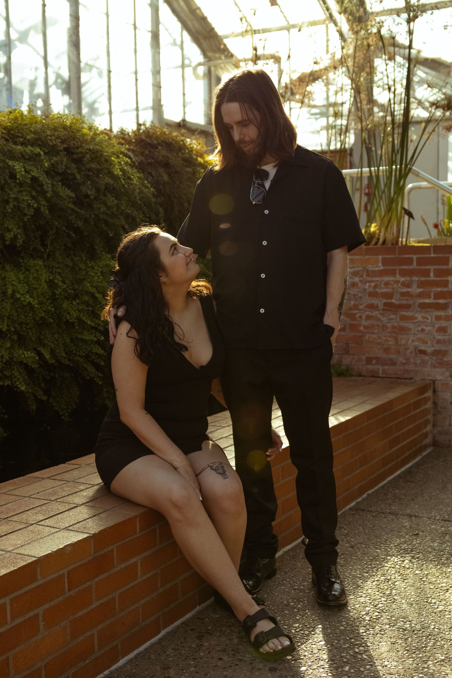 Couple in black outfits posing in a sunlit greenhouse, woman sitting on brick ledge looking up at long-haired man.