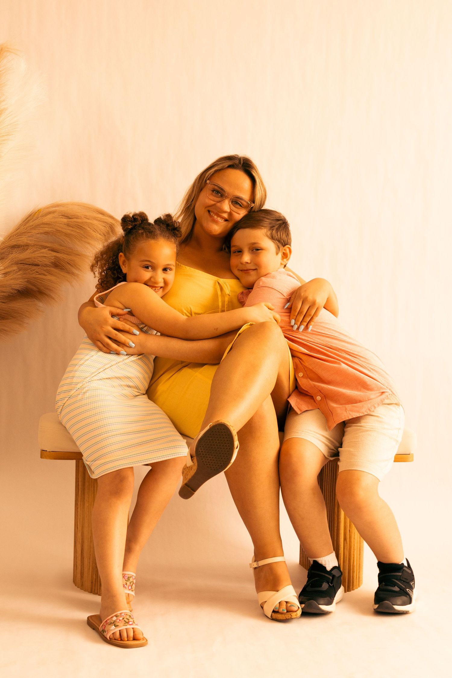 A woman in a yellow dress sits on a bench, hugging two smiling children in a warm, softly lit studio setting.
