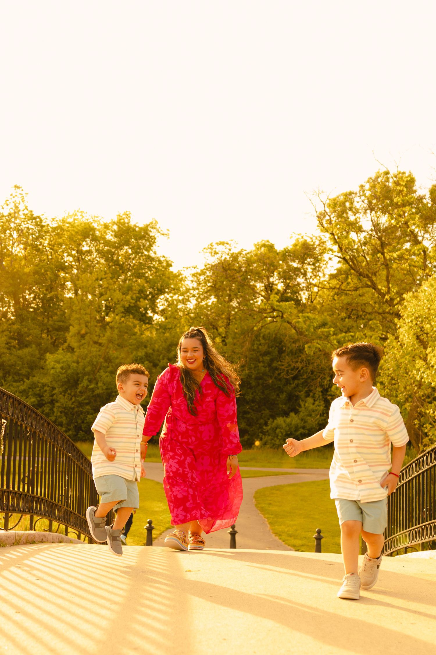 A joyful mother in a red dress runs with her two young sons on a sunlit park bridge surrounded by lush green trees.