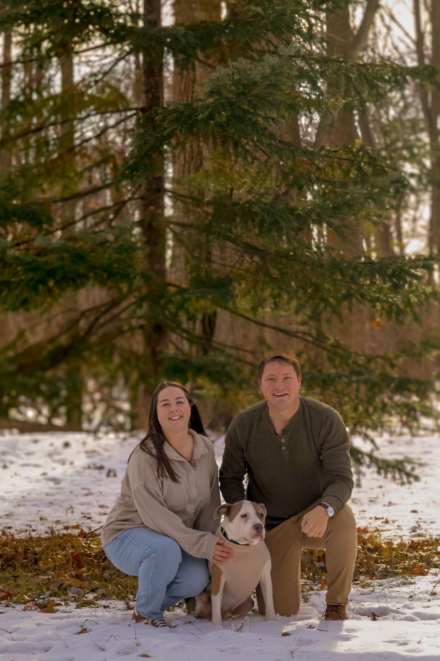 A smiling couple crouches in snow with their pit bull dog in front of evergreen trees in a winter forest.