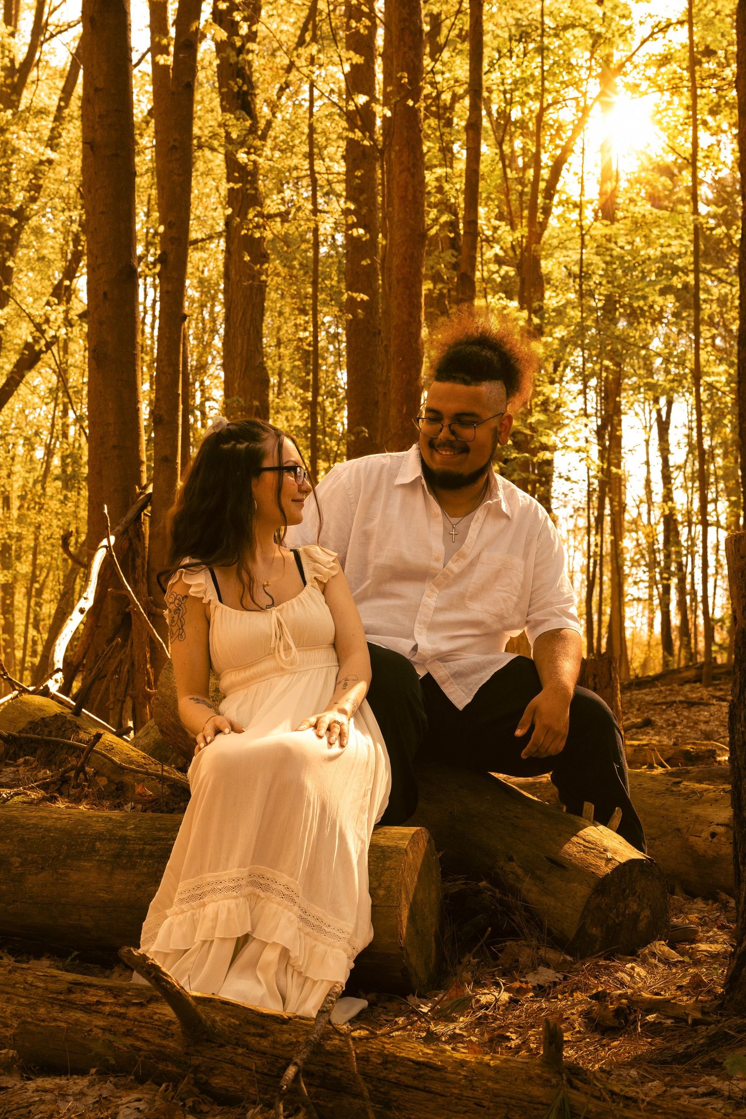 Couple sitting on fallen logs in golden sunlit forest, smiling at each other in casual elegant attire.