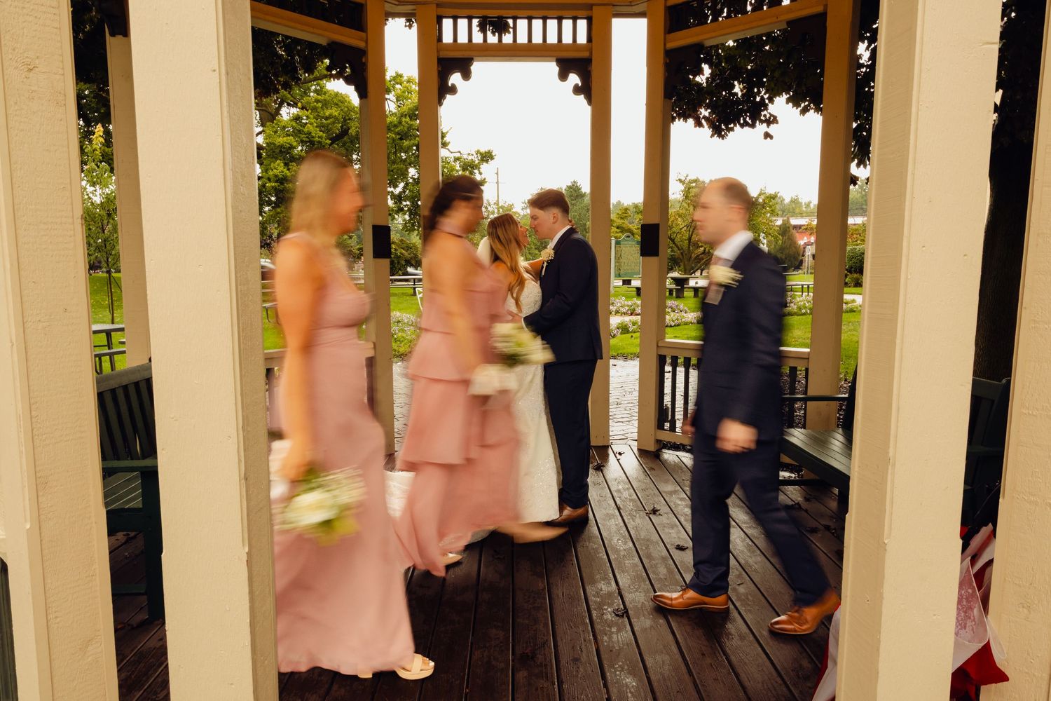 Bridesmaids in blush pink dresses and groomsmen in navy suits walk across a wooden gazebo deck during a wedding party celebration.