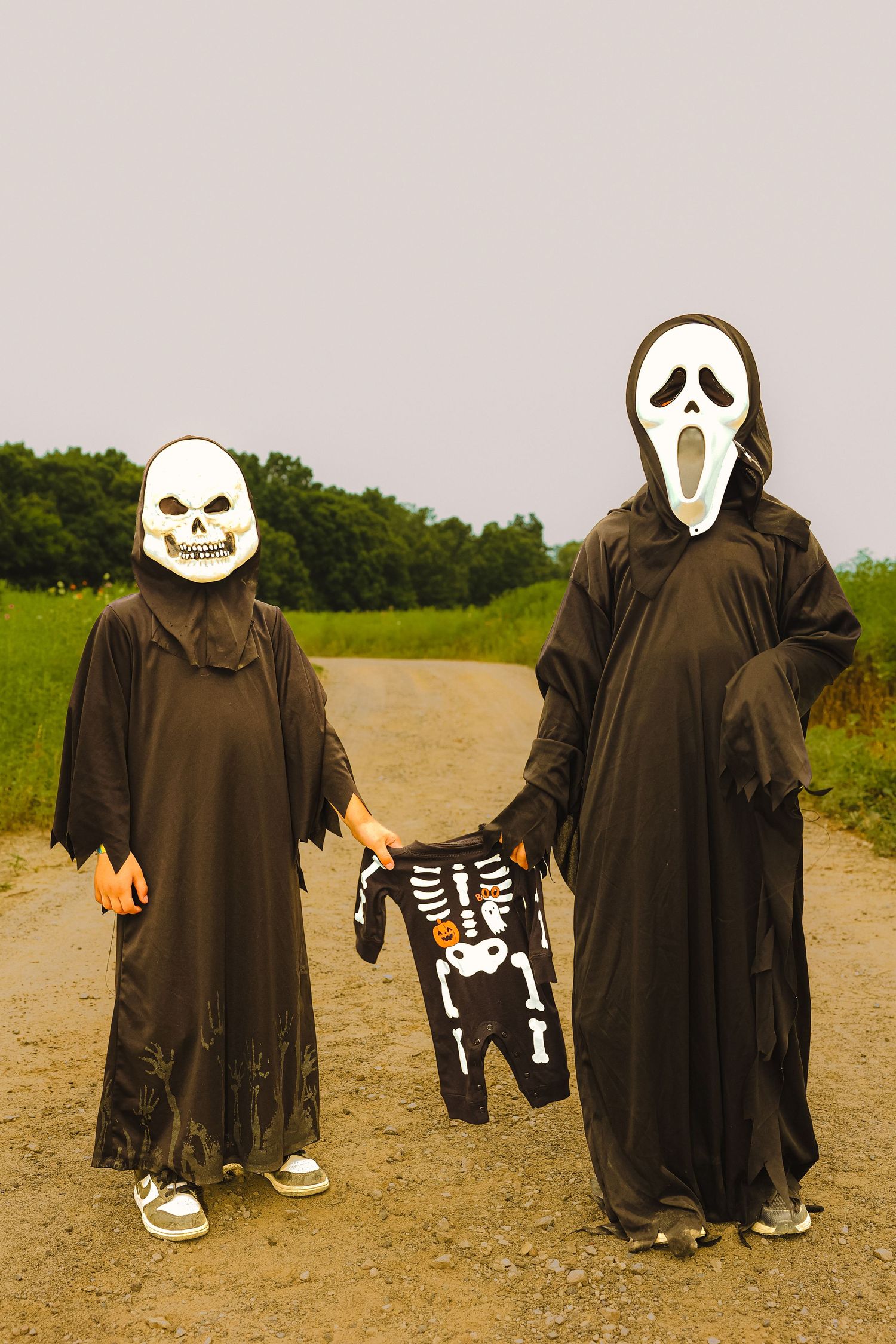 Two children in black Halloween costumes with skull and Scream masks stand on a dirt path holding a skeleton outfit.