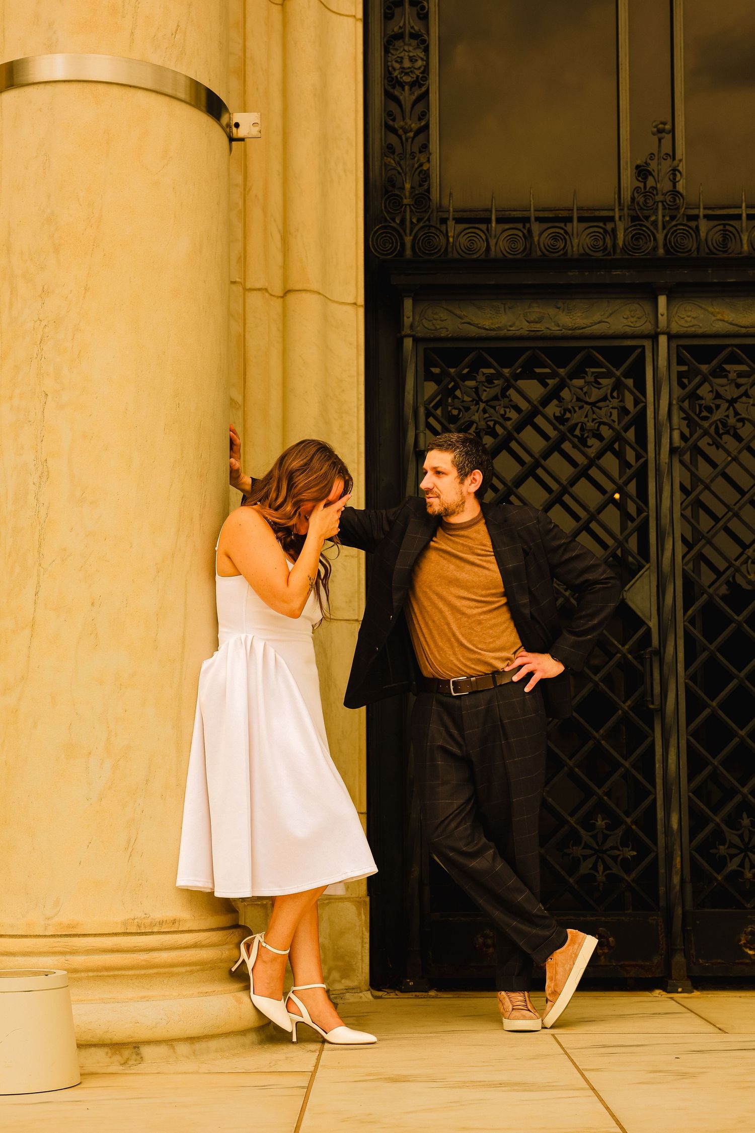 Couple in elegant attire posing by ornate building columns, woman in white dress and man in dark blazer.