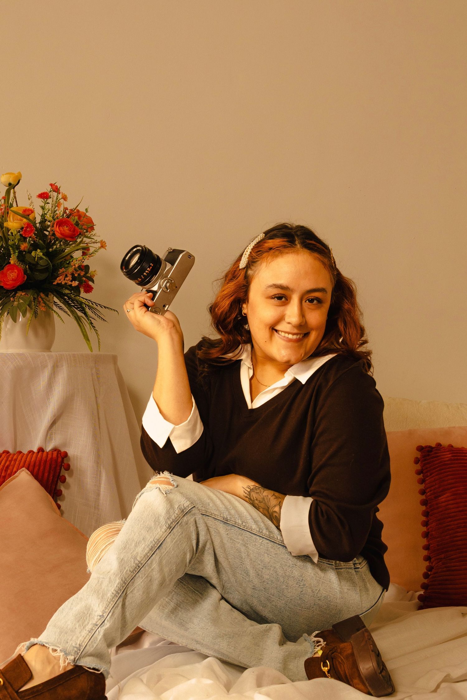 Smiling young woman holding a vintage camera, sitting on a bed with colorful flowers, wearing jeans and black sweater.