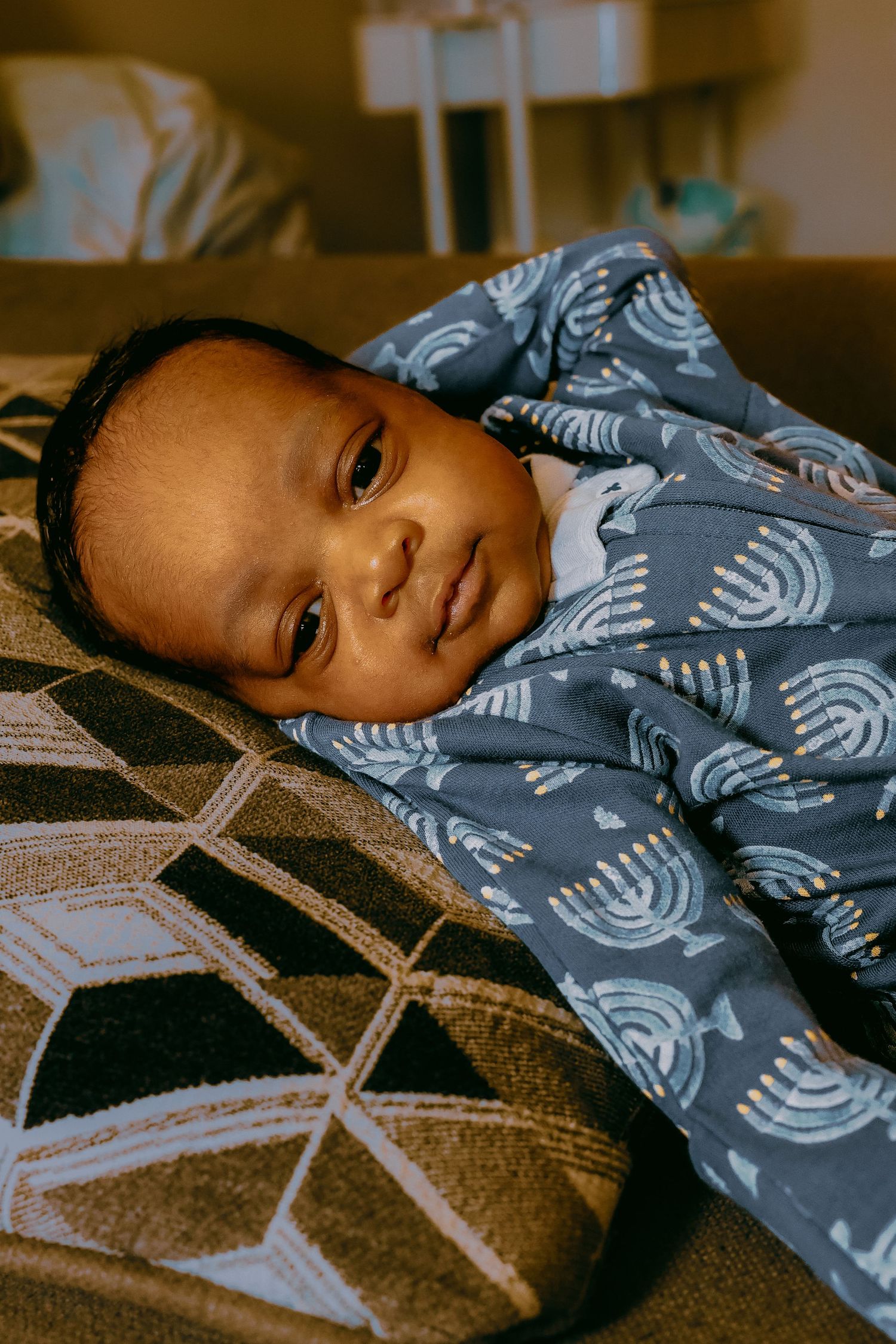 Newborn baby in blue Hanukkah menorah pajamas lying on a patterned blanket on a couch.