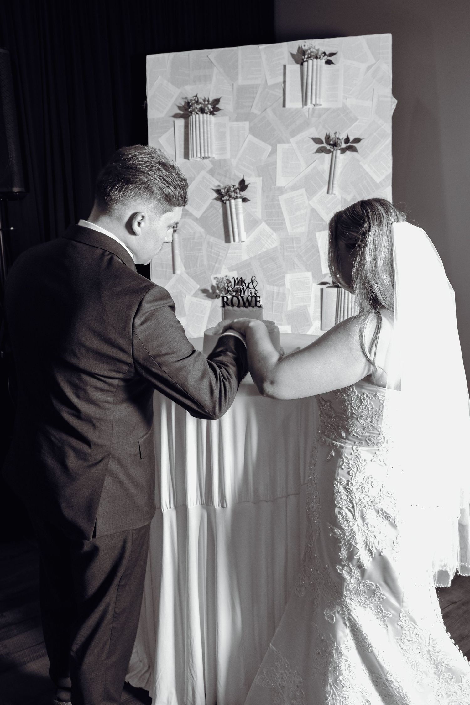 Black and white photo of a bride and groom from behind standing at a draped table with a cake topper before a book page wall display.
