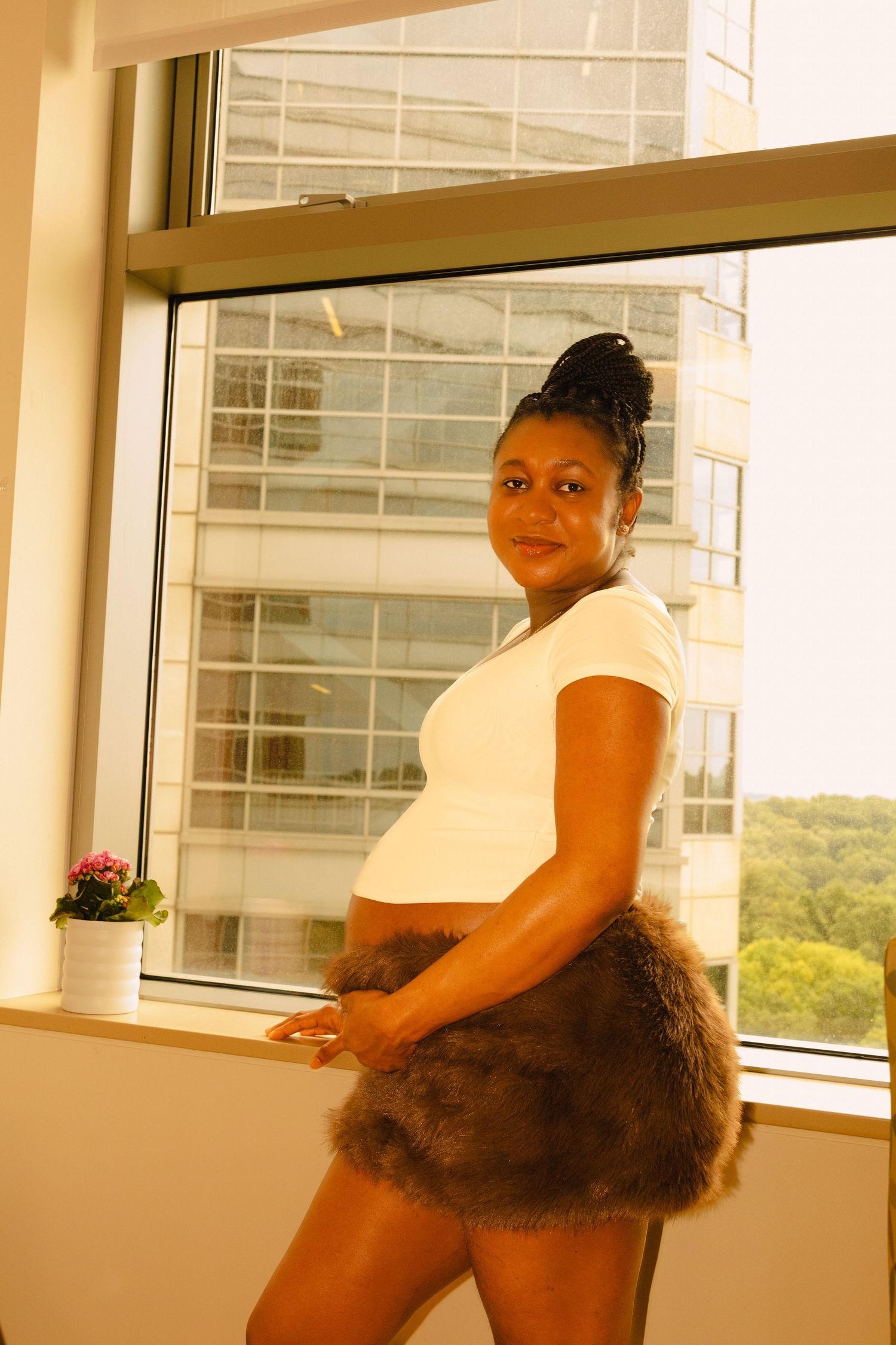 Pregnant woman in white top and fur skirt posing by large window with city building view.