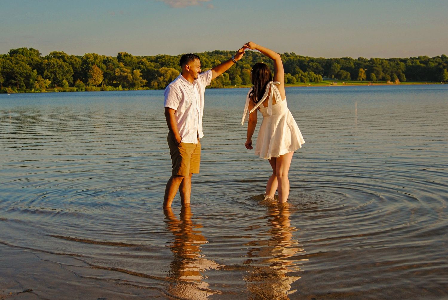 Two people wade in calm lake waters at sunset while casting long shadows across the rippling surface.