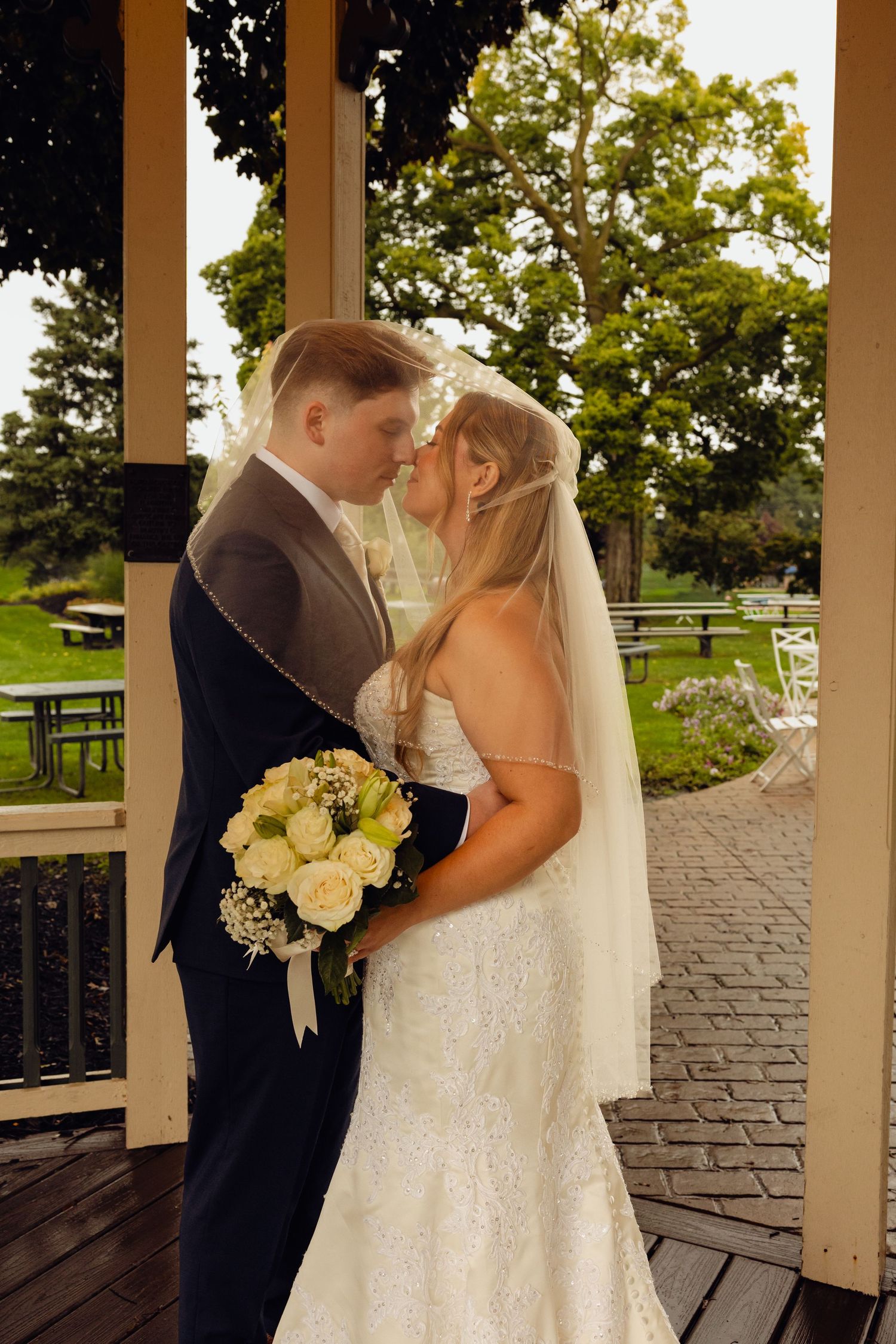 A bride and groom share an intimate moment under a veil in a park gazebo, holding a bouquet of white roses.