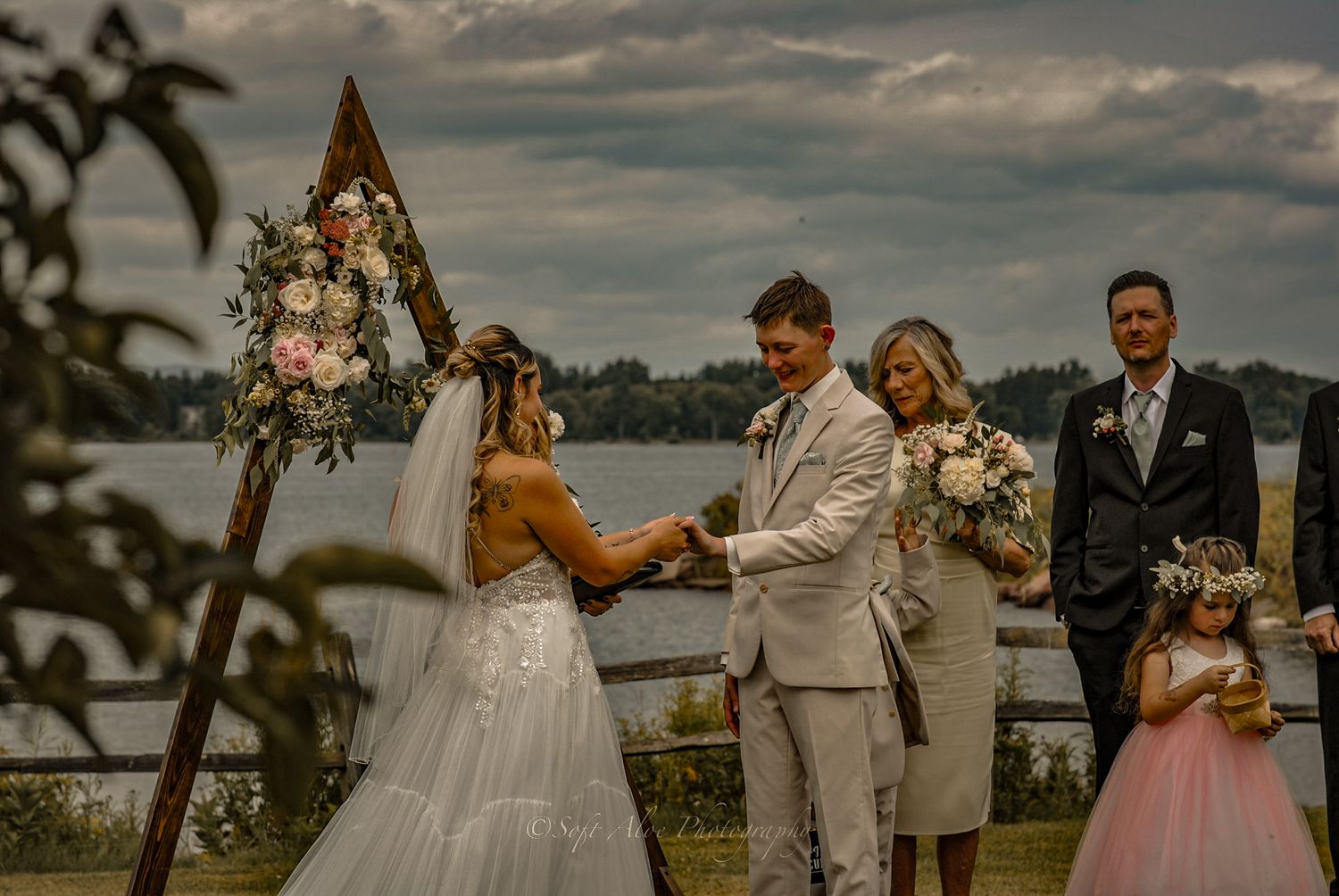 A lakeside wedding ceremony under a triangular floral arch with guests gathered on a cloudy day.