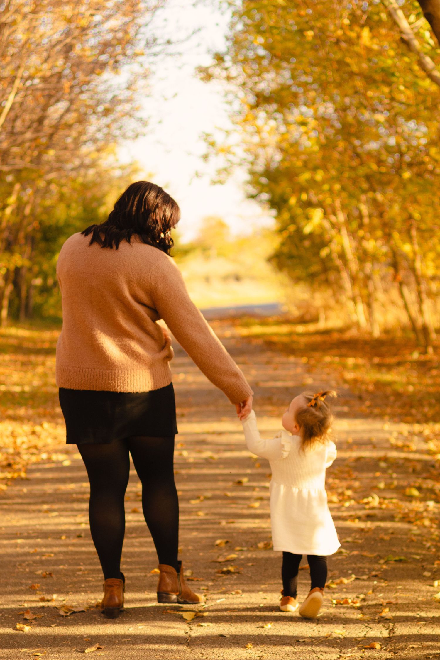 Mother and toddler daughter holding hands walking on a leaf-covered autumn park path surrounded by golden yellow trees.