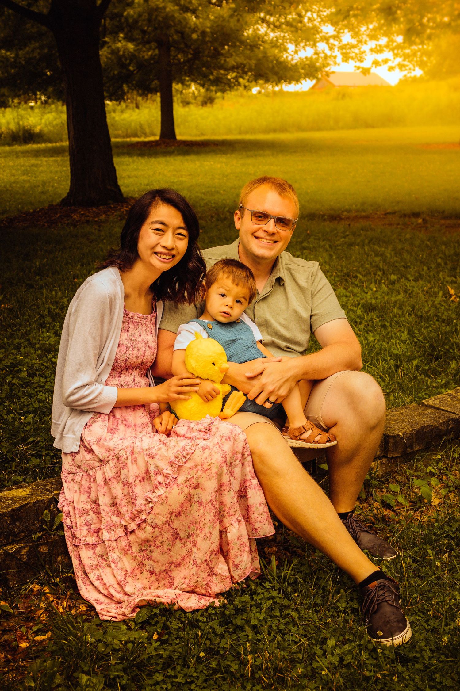 Family portrait of couple with toddler holding yellow stuffed animal, sitting on grass at golden hour sunset in park.