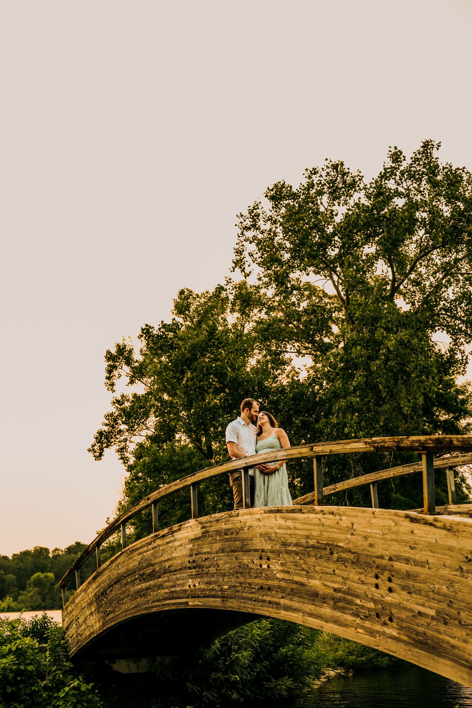 Couple sharing a romantic kiss on a wooden arched bridge surrounded by lush green trees at golden hour.