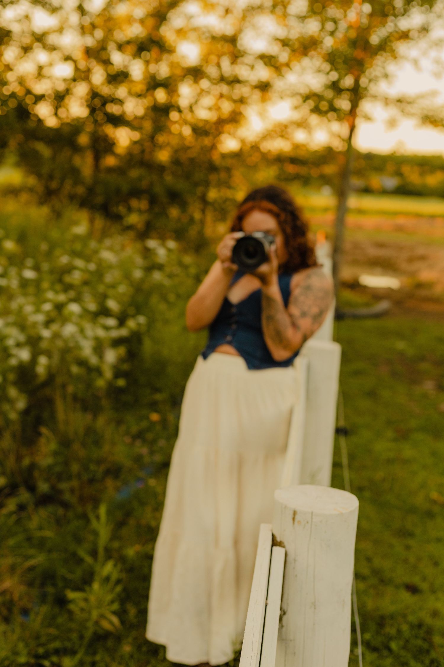Woman in white skirt and blue top photographs with camera outdoors near white fence at golden hour.