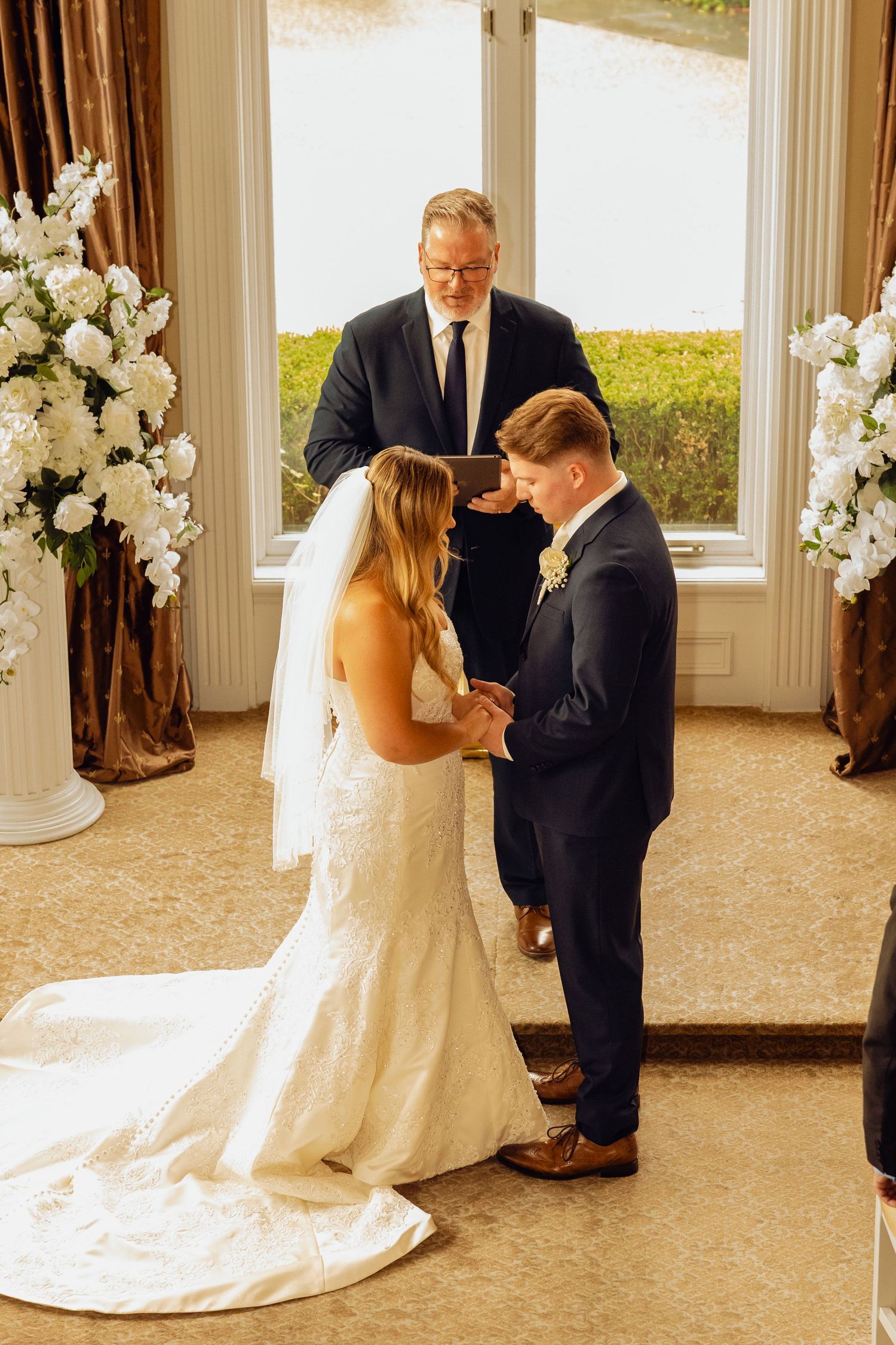 Bride in a white lace gown with long veil and groom in a navy suit exchange vows as an officiant looks on during an indoor wedding ceremony.