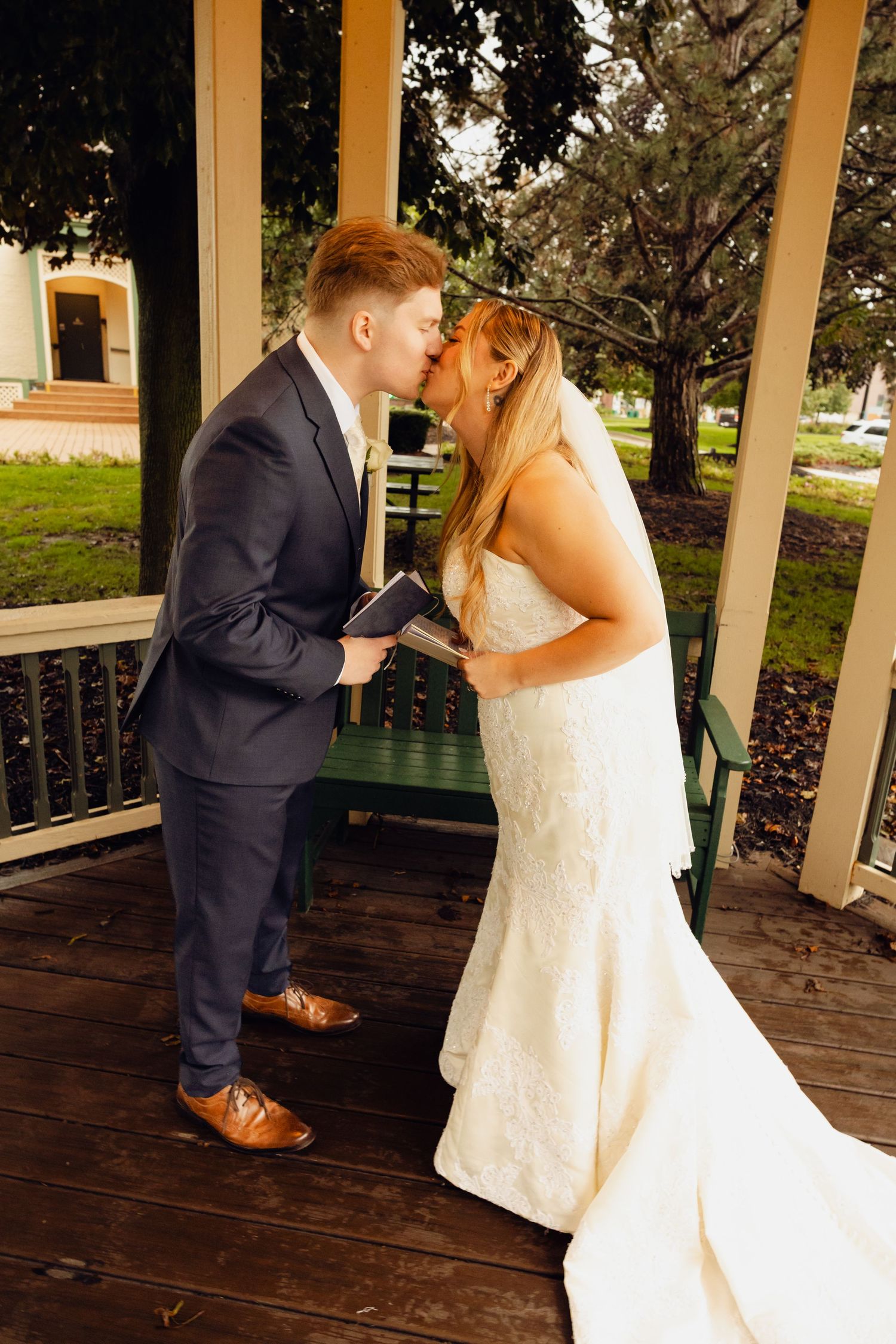 A bride in a lace wedding gown and a groom in a navy suit share a kiss while holding books in a wooden gazebo.