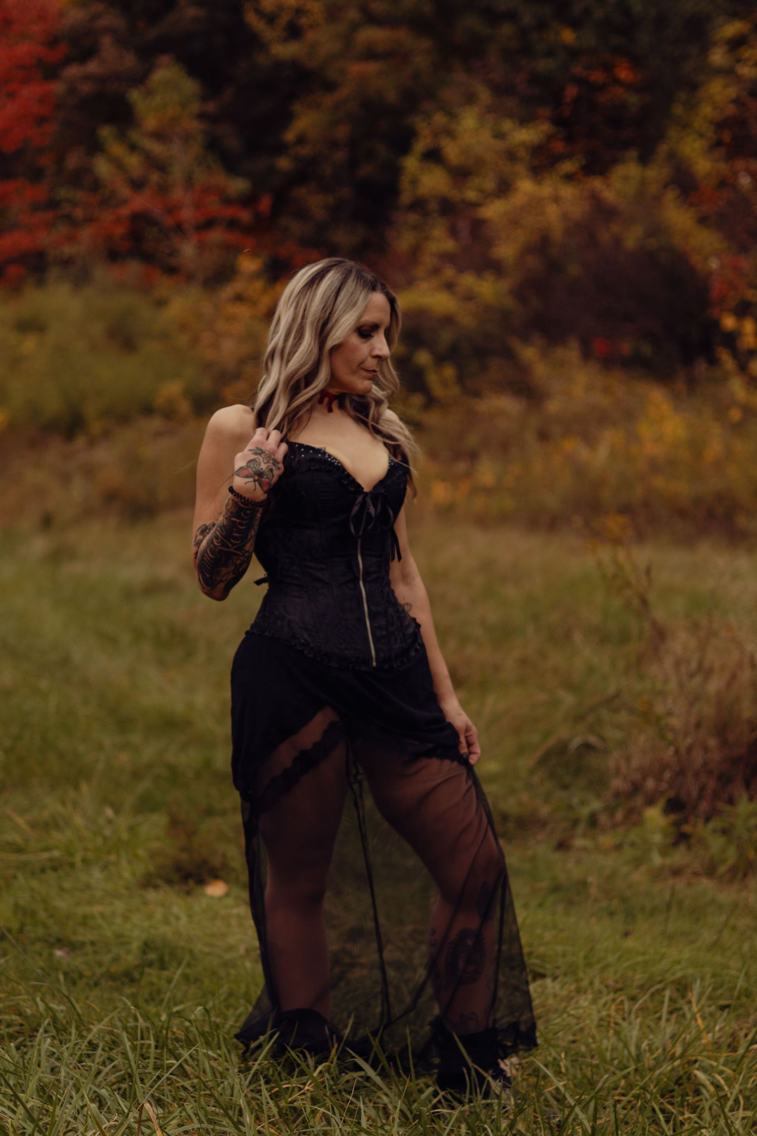 Woman in black sheer lace gown posing outdoors in autumn field with colorful fall foliage background.