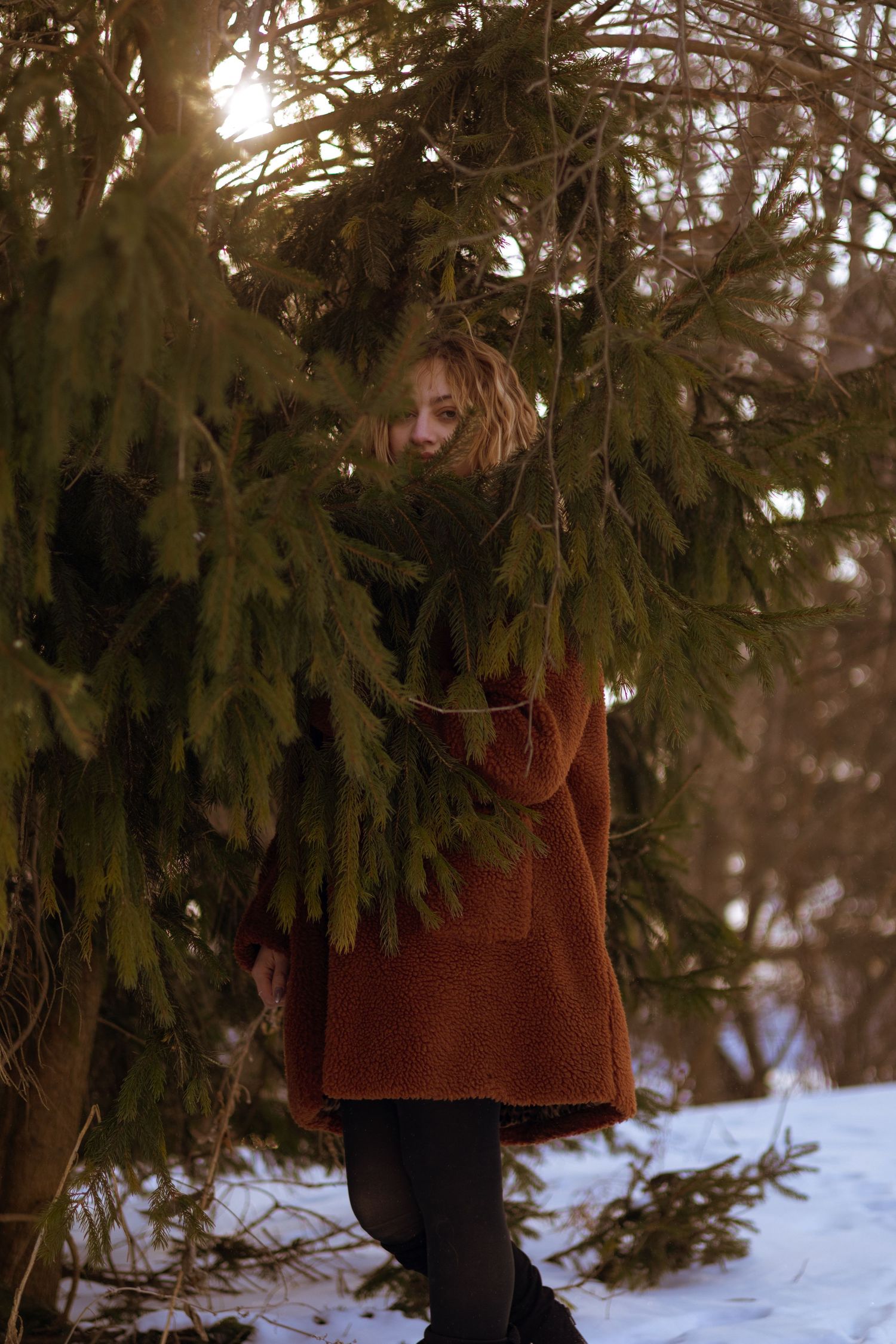 Woman in rust sweater standing among snow-dusted evergreen branches in a winter forest at golden hour.