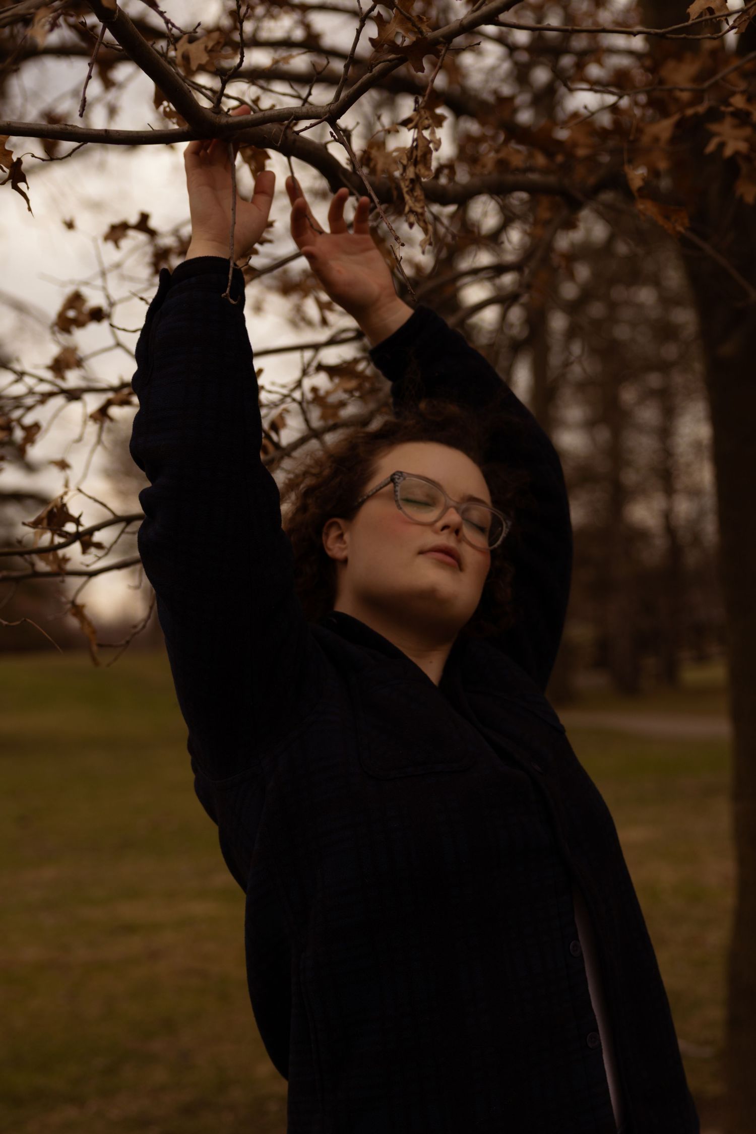 A young woman in a black coat and glasses reaches up to touch bare autumn tree branches in a moody outdoor park setting.