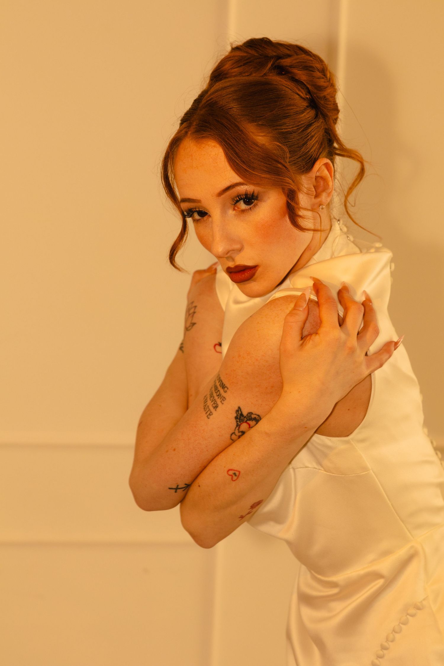 Young woman in white dress with tattoos on arms, red lips, updo hairstyle, posing against a warm beige wall.