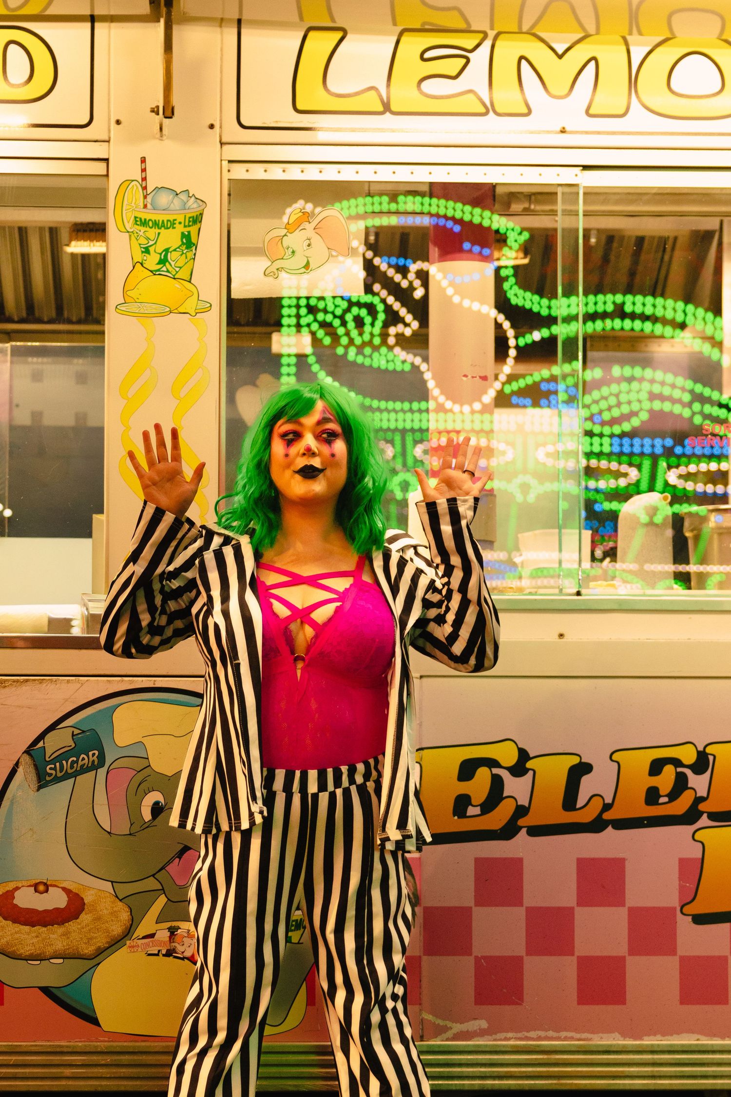 Person in green wig and black-and-white striped suit poses dramatically at a colorful carnival lemonade stand at night.