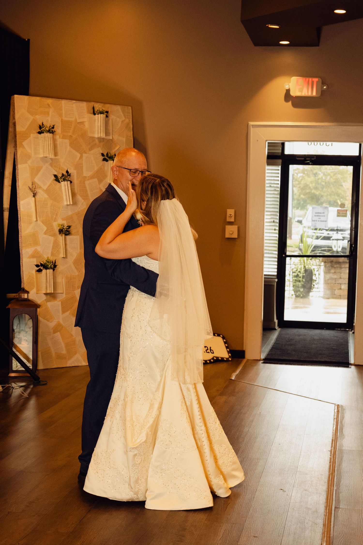 A bride in a white gown and veil shares a tender dance with a man in a navy suit in a modern venue with a wooden floor.