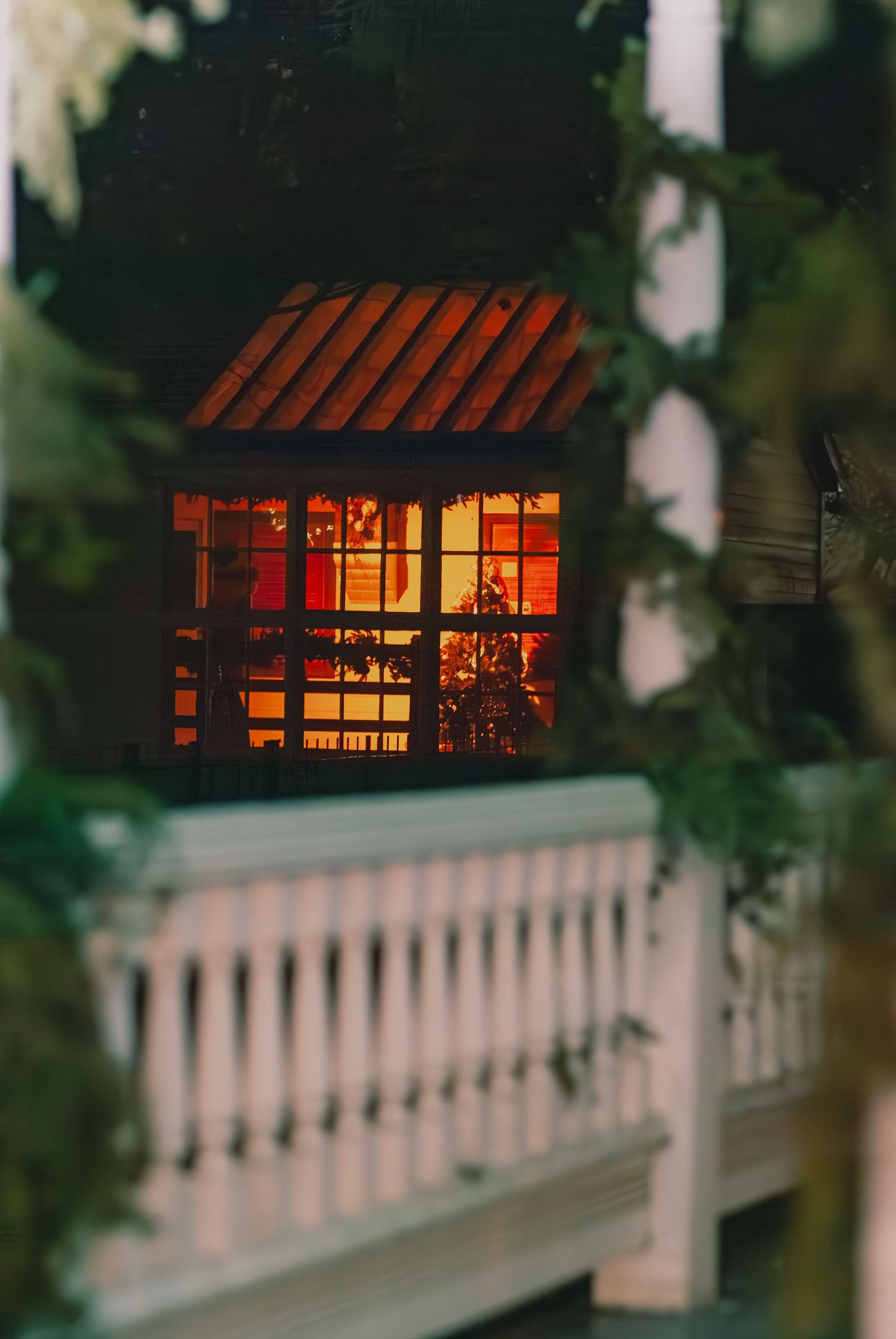 Glowing red windows of a structure behind white railings, framed by tree branches at dusk.